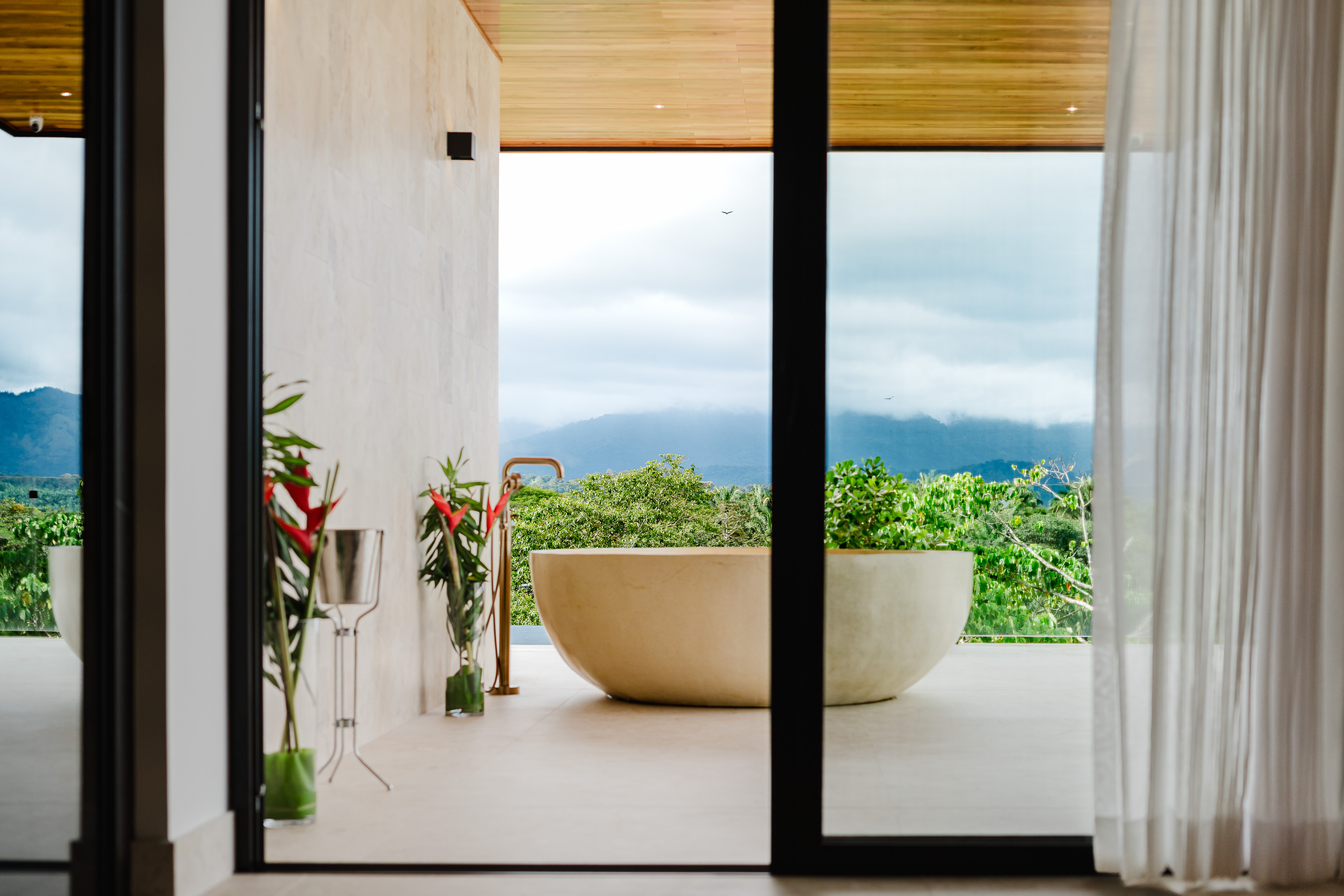 An open-air bathing area features a freestanding tub on a terrace, overlooking forested mountains and the sky through large glass doors.