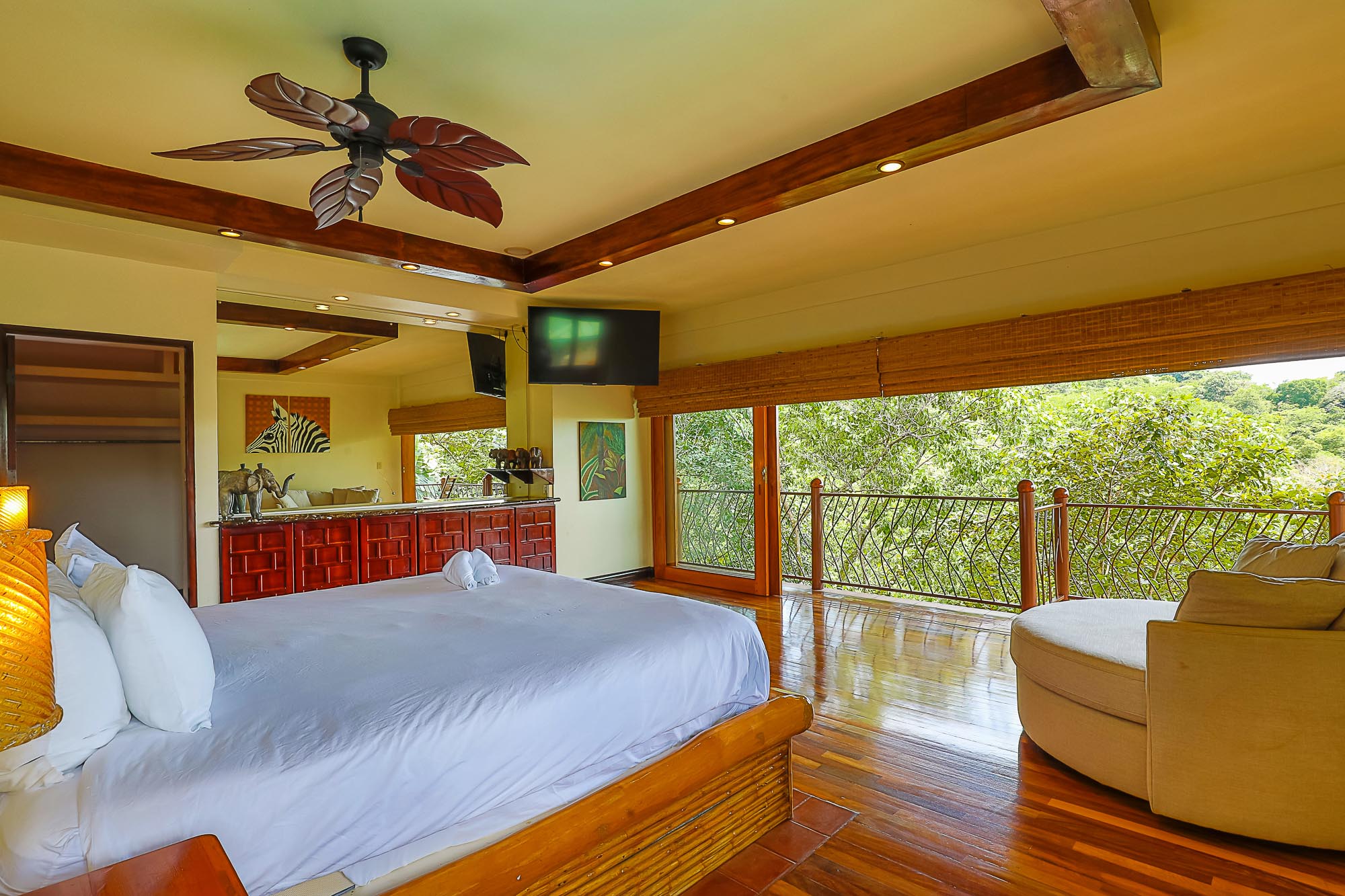 This bedroom offers a large bed, polished wood floors, and floor-to-ceiling glass doors that open to a private balcony overlooking the rainforest, providing natural light and an open-air feel.