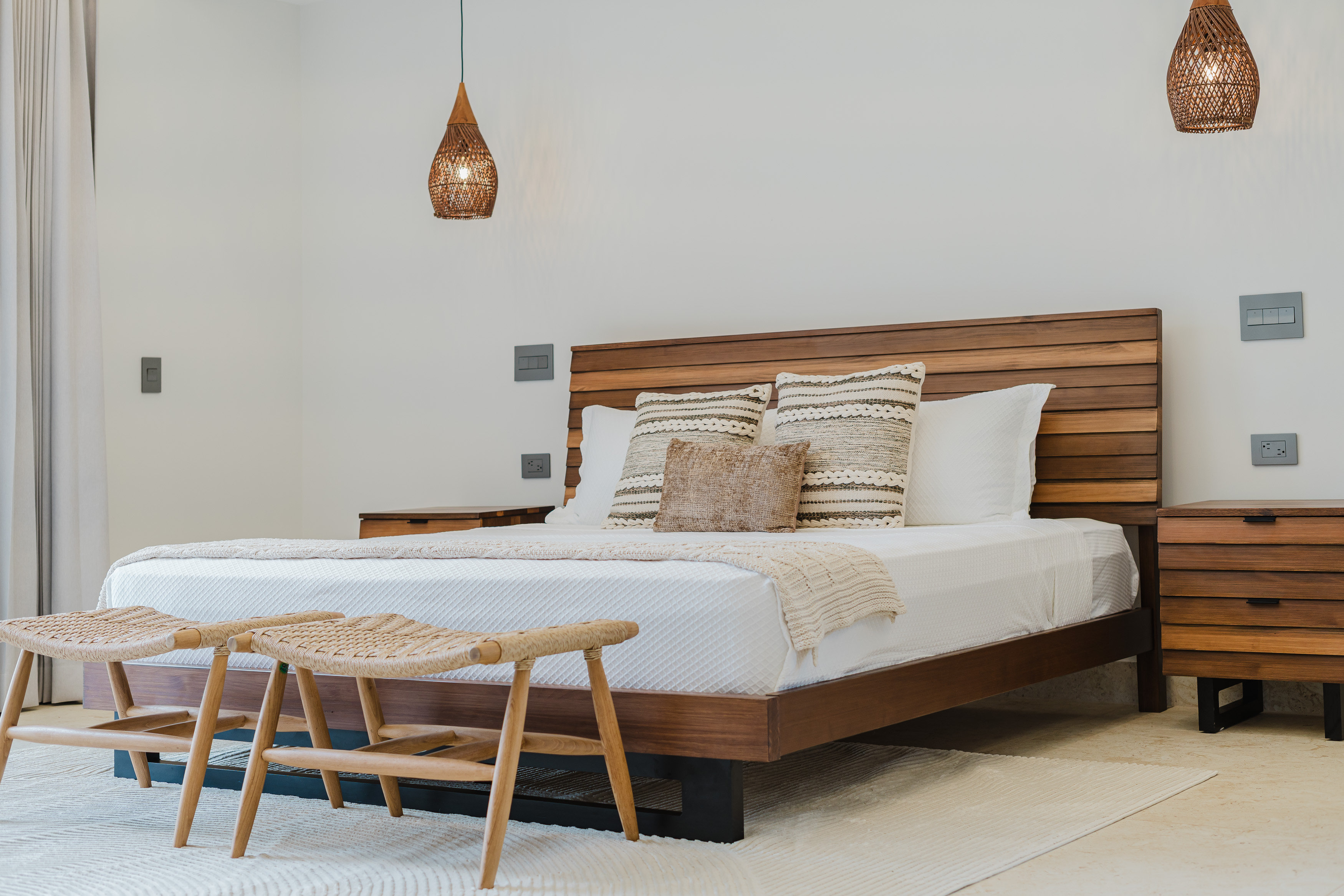 A bright bedroom with a slatted wooden bed frame and headboard, matching nightstands, and woven pendant lights, illuminated by natural light from a curtained opening.