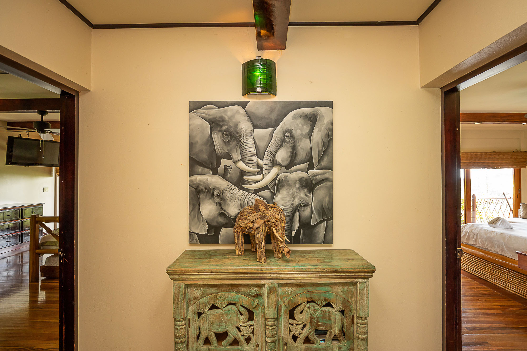 A welcoming hallway featuring locally-sourced wood accents, a carved console, and elephant-themed artwork connects to a bright, natural-wood-furnished bedroom and a comfortable living area.