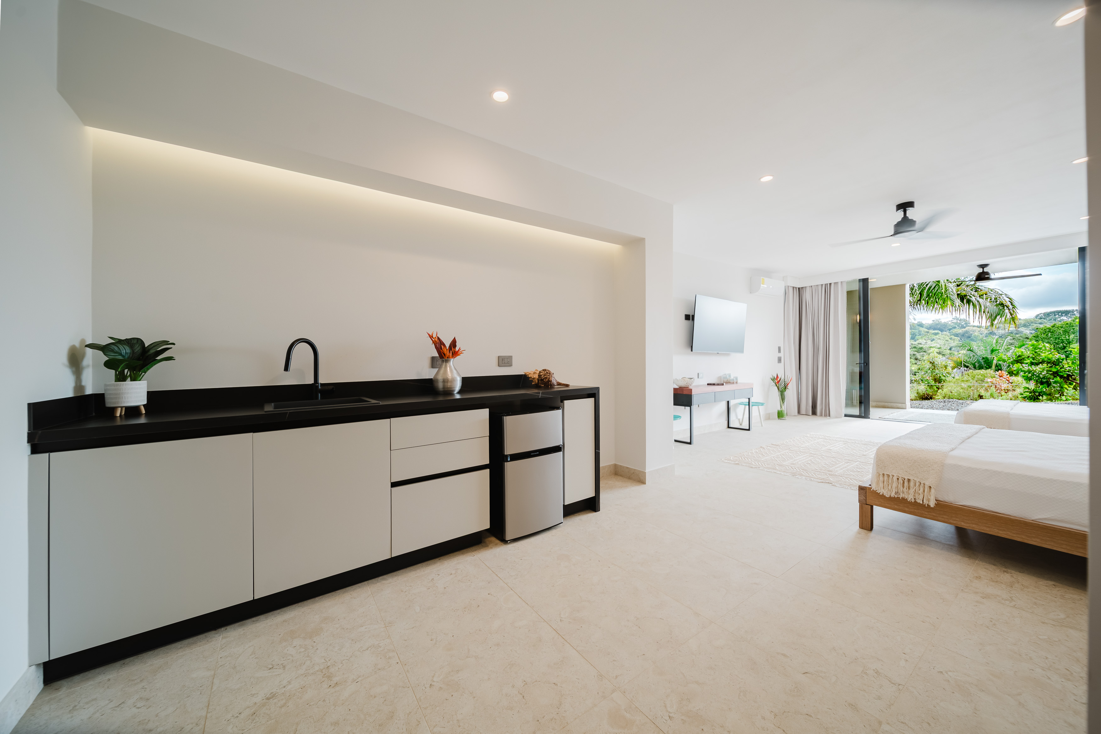 This bright bedroom suite includes two beds and a small kitchenette, with wide glass doors opening to a lush tropical garden and ample natural light.