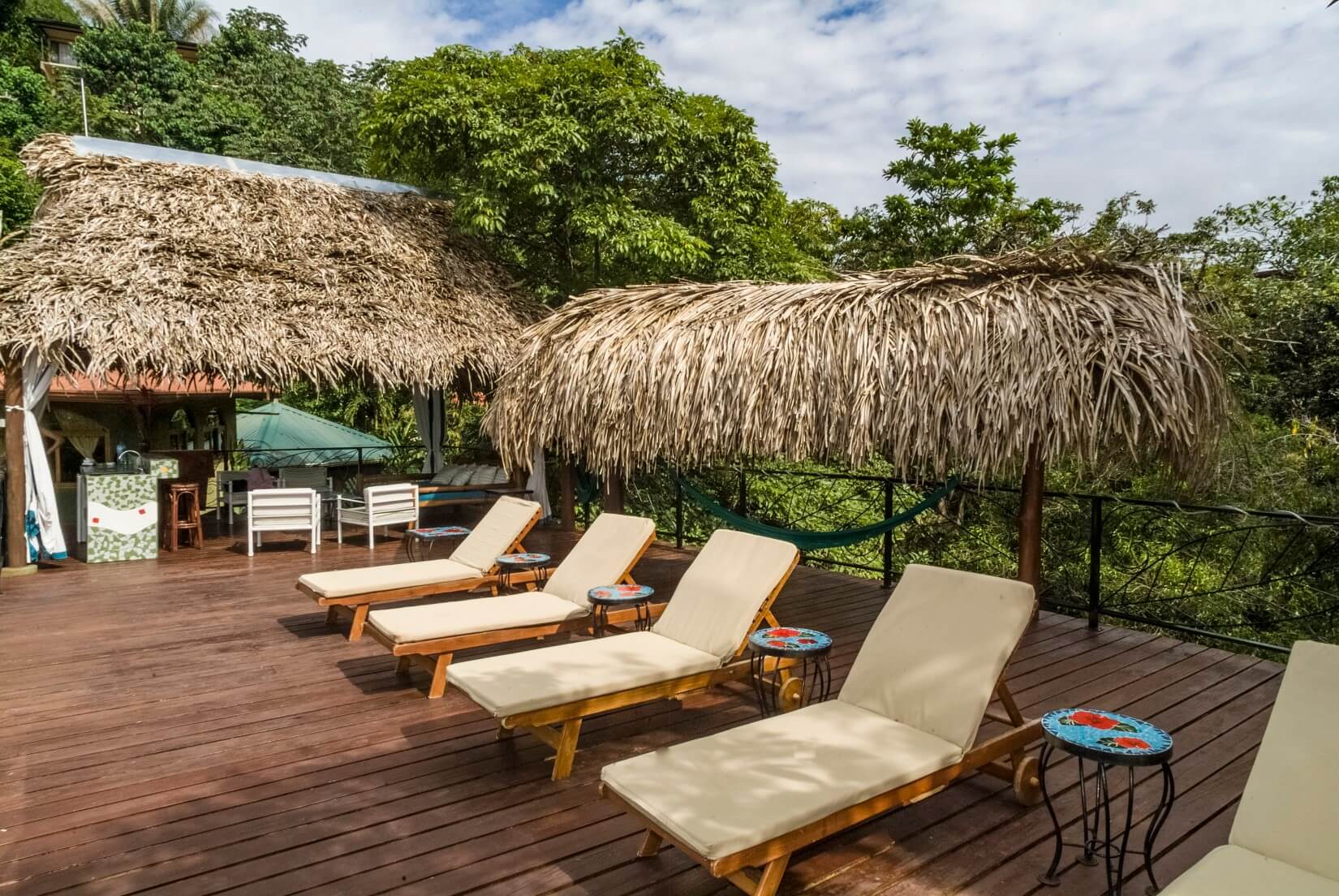 A spacious wooden deck with lounge chairs and thatched palapas includes a wet bar, opening to views of the surrounding tropical jungle under natural light.