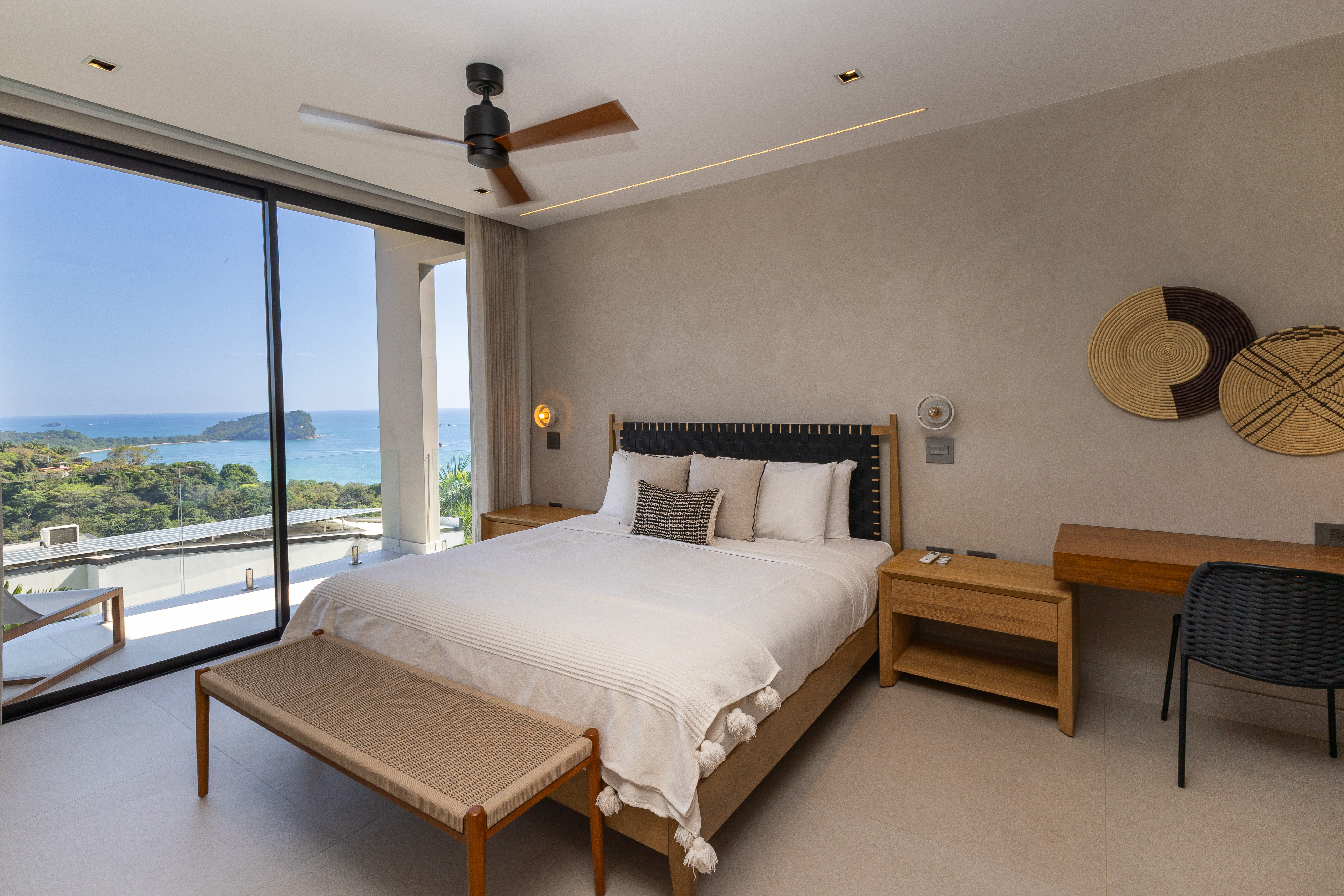 A spacious bedroom with a large bed and full-height glass doors offers an expansive view of the Pacific Ocean and jungle, filling the contemporary room with natural light.