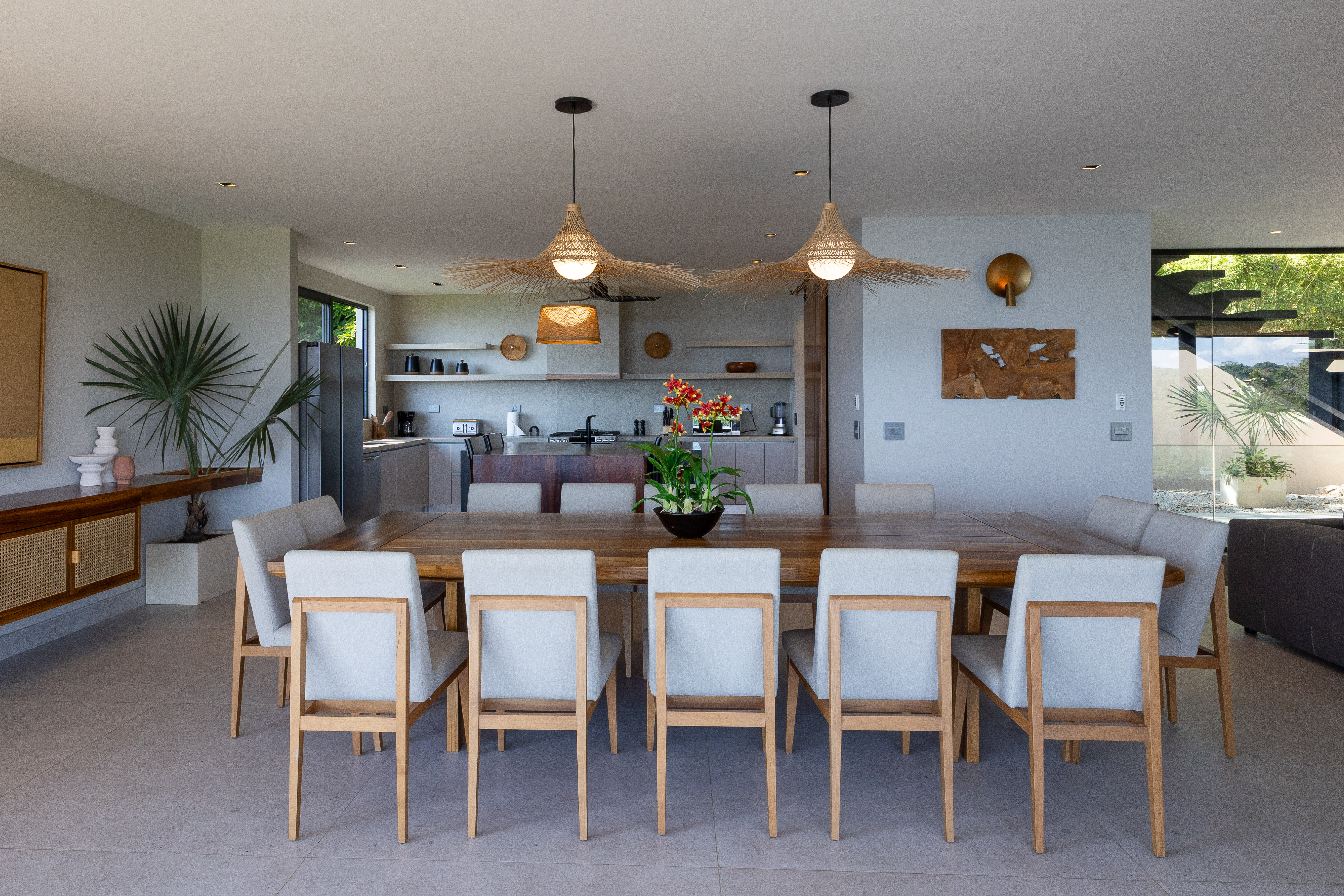 The dining area features a long table with upholstered chairs, an open connection to the kitchen, and natural light from glass walls offering views of surrounding jungle greenery.