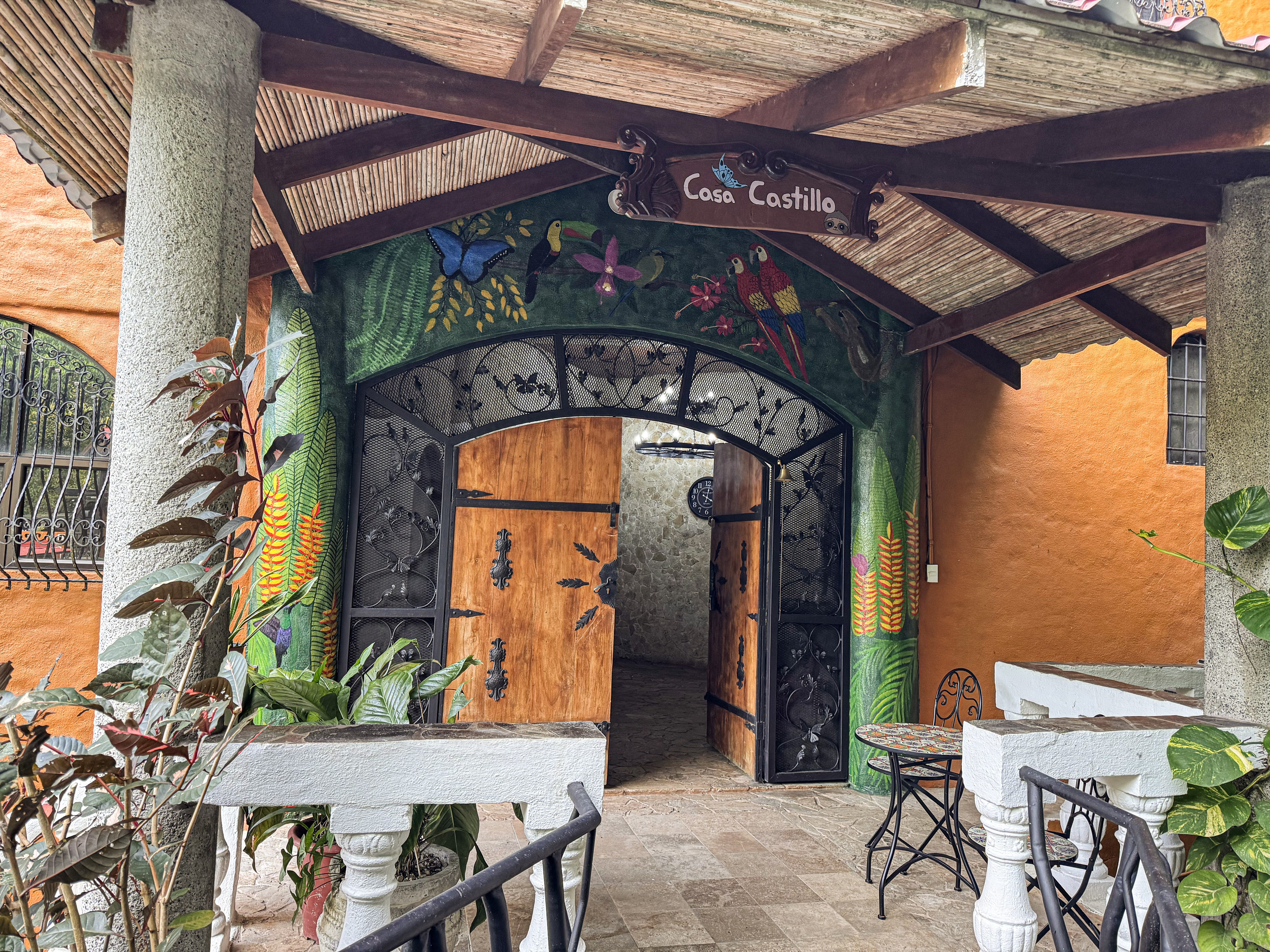 The villa's entry features richly painted walls adorned with tropical murals, substantial carved wooden double doors framed by custom wrought iron, and an inviting slatted wood ceiling.