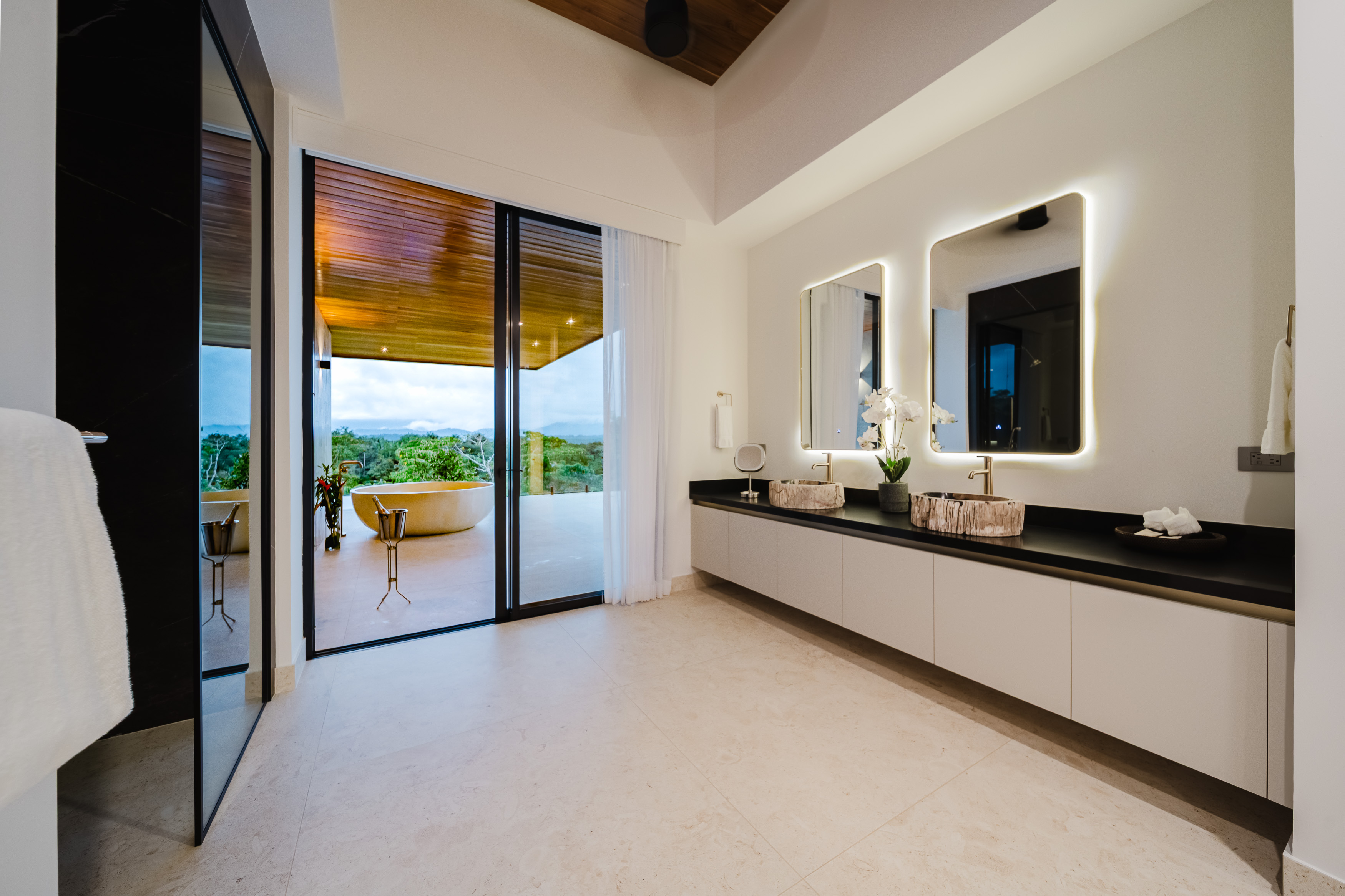 The spacious bathroom features a double vanity and opens through large glass doors to a private outdoor terrace with a freestanding tub overlooking a lush jungle landscape.