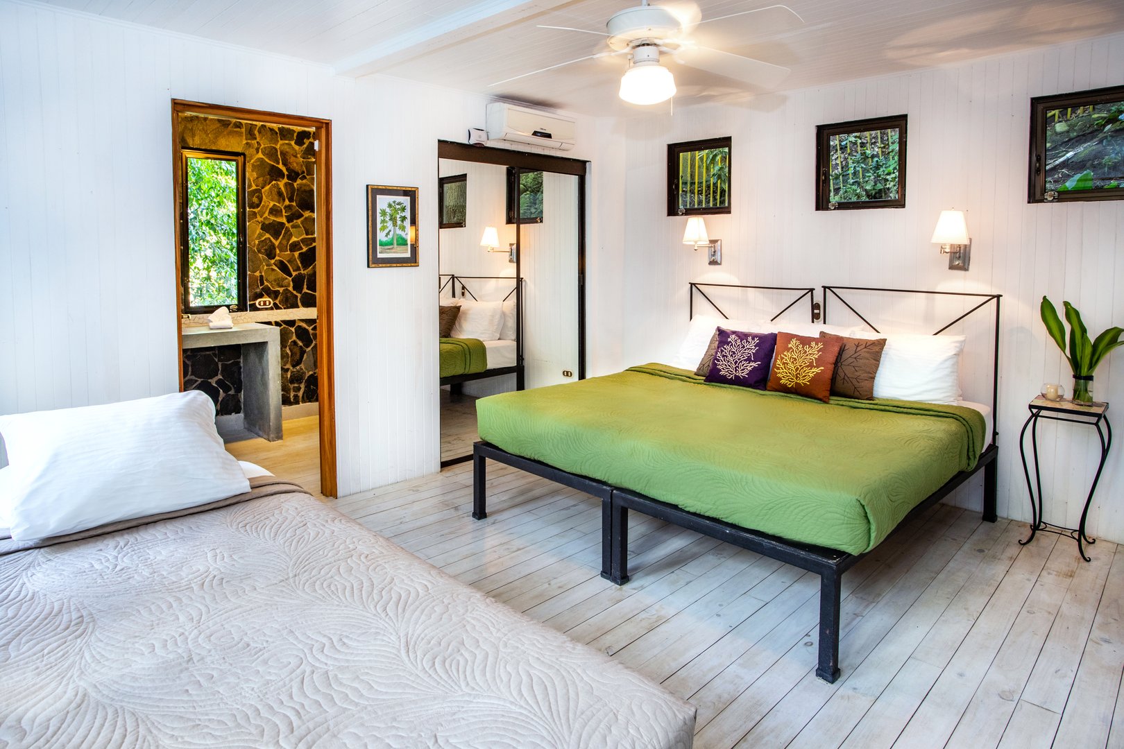 This bright bedroom with white paneled walls and light wood floors provides two beds, tropical jungle views, and an en-suite bathroom.