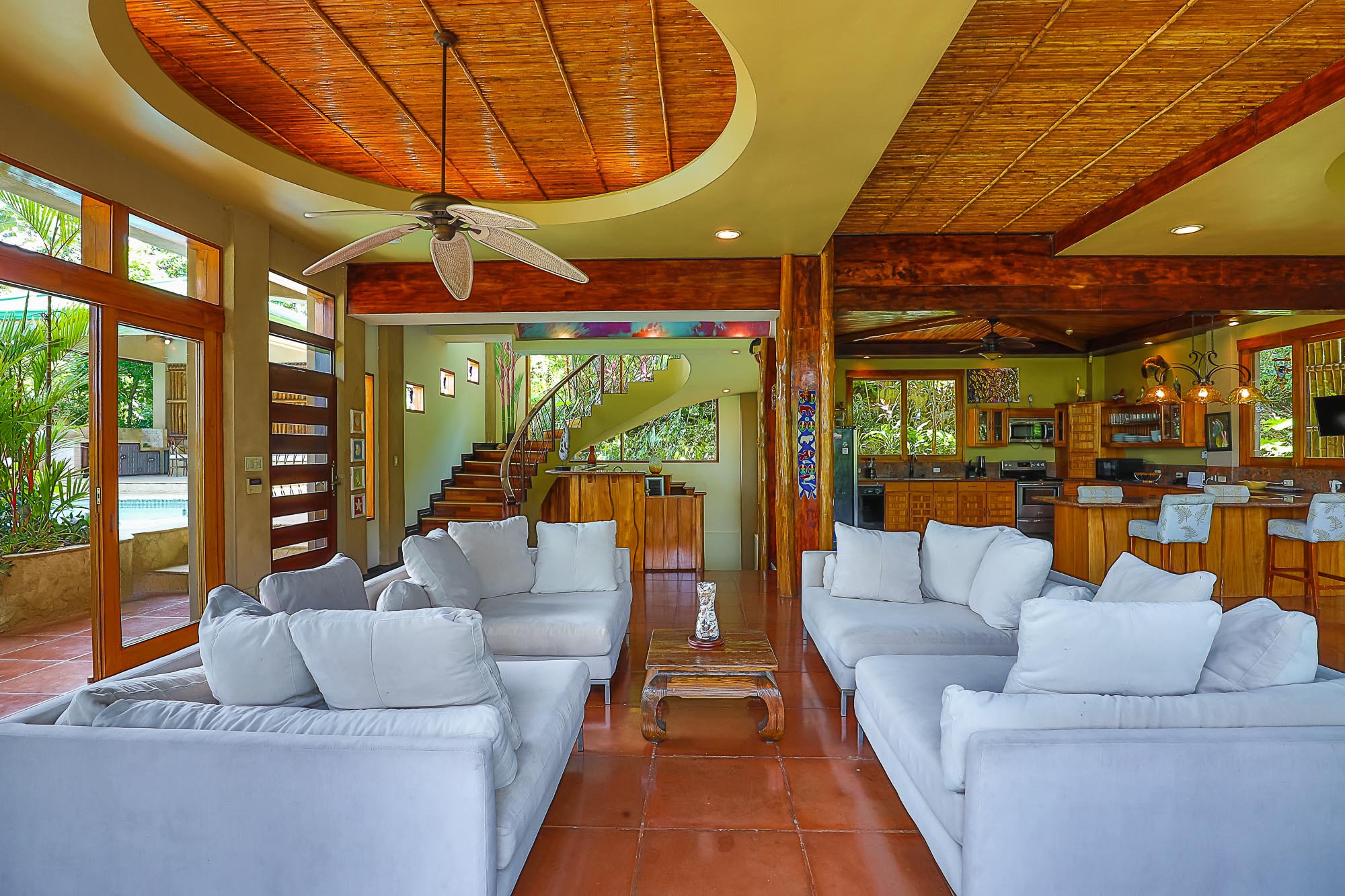 An open-plan living room with comfortable white sofas features a high bamboo ceiling and direct access to a pool terrace, with a full kitchen and a spiral staircase visible within the tropical-view space.