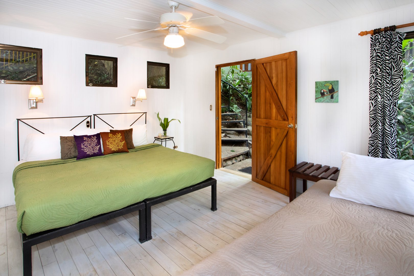 This bright bedroom with white paneled walls and flexible bedding options features jungle views from windows and an open wooden door leading to stone steps.