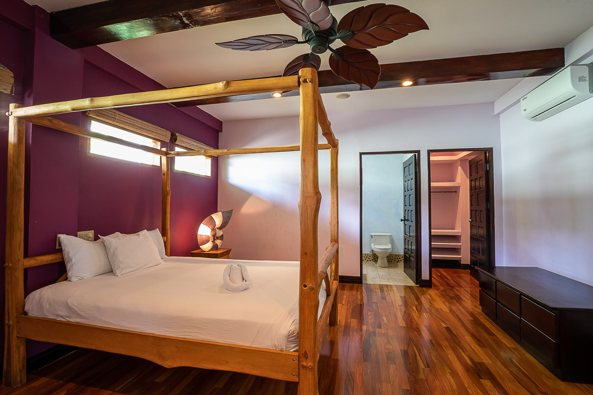 This bedroom features a natural wood four-poster bed, polished wood floors, exposed ceiling beams, and receives natural light from high windows, with an adjacent en-suite bathroom and closet.