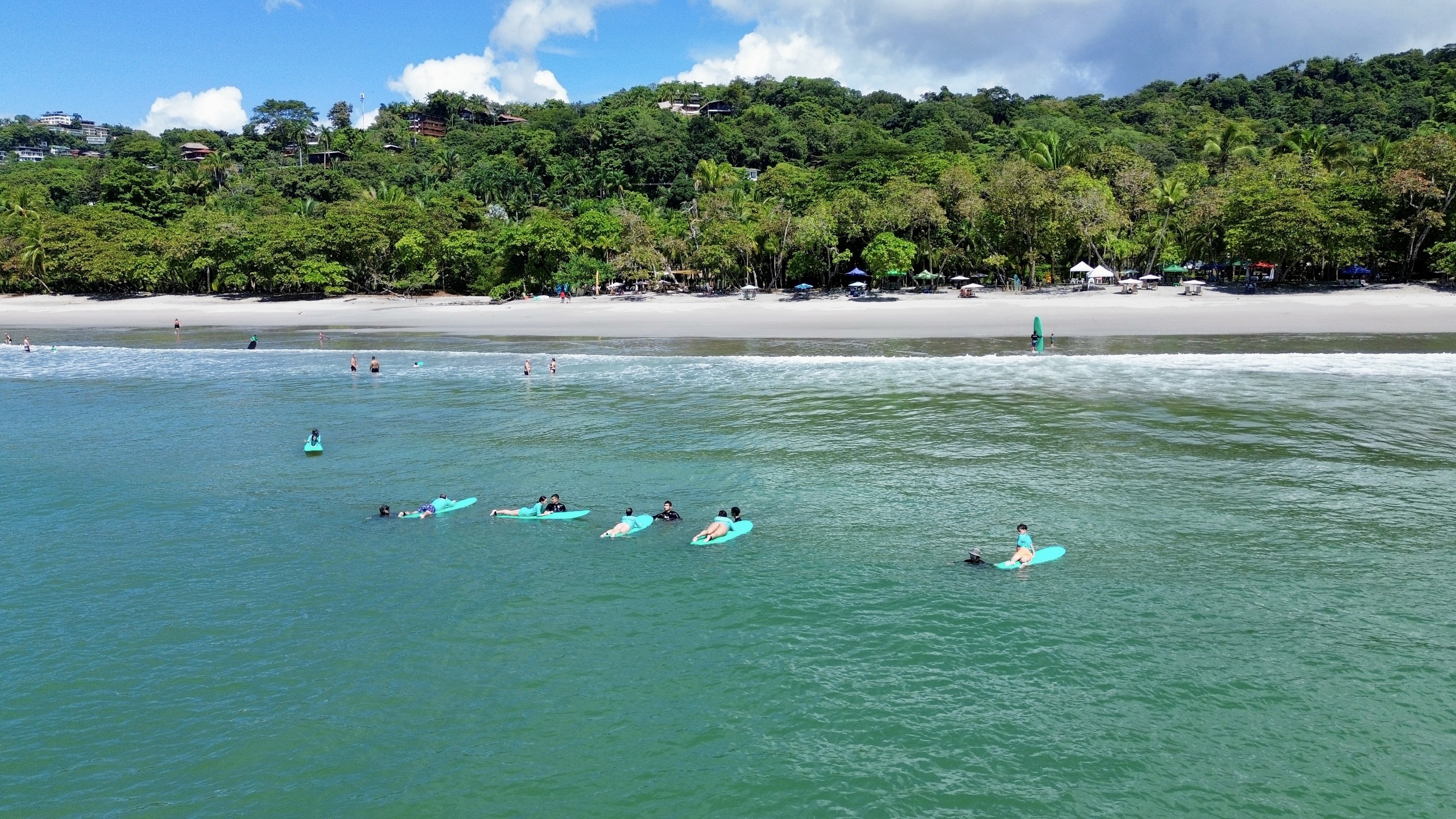 A wide aerial view shows the turquoise ocean with people on surfboards, a sandy beach with umbrellas, and a verdant hillside dotted with buildings under a bright blue sky.