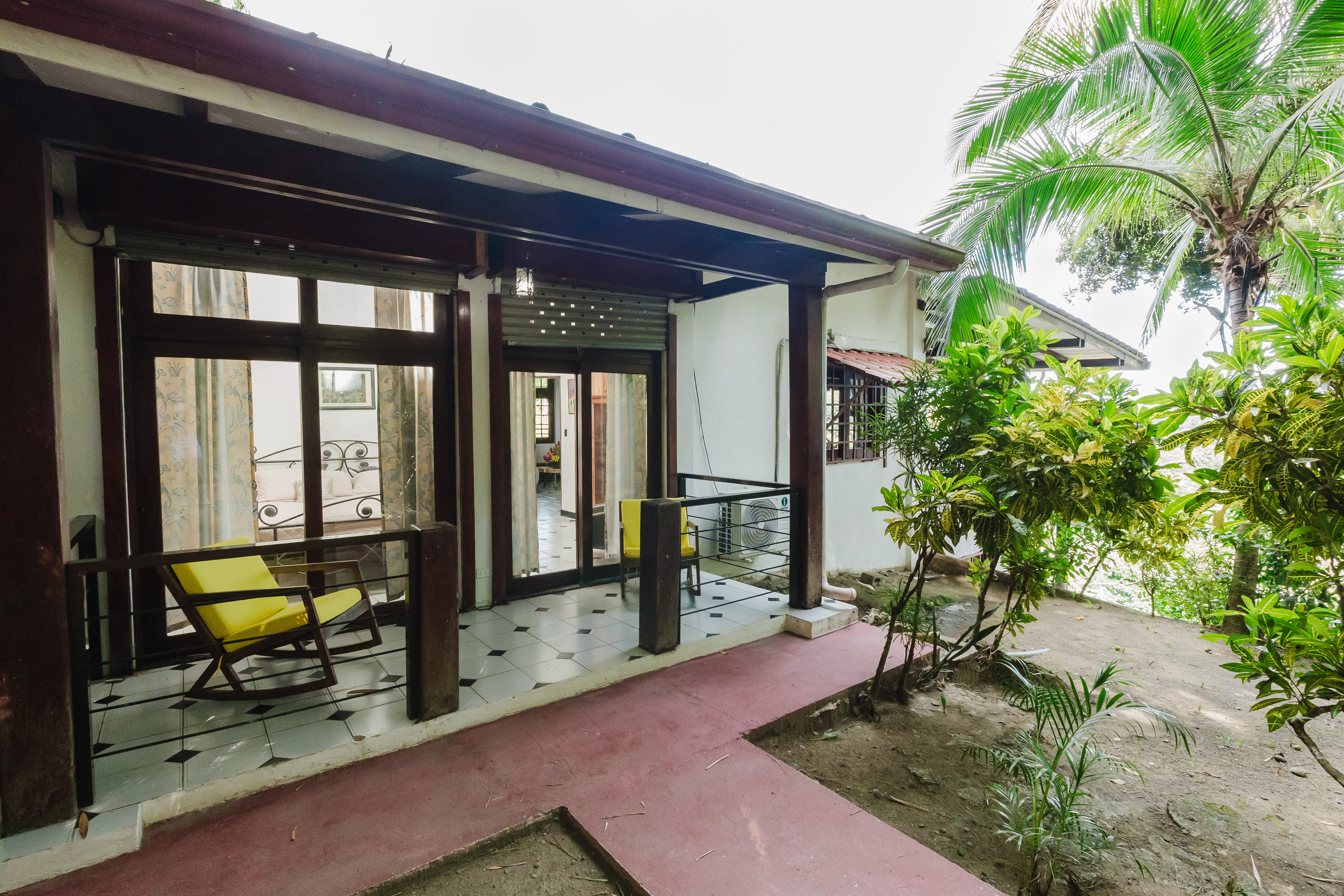 A bright, covered outdoor terrace with dark wood columns and tiled flooring provides an inviting seating area, overlooking a lush tropical garden with a large palm tree.