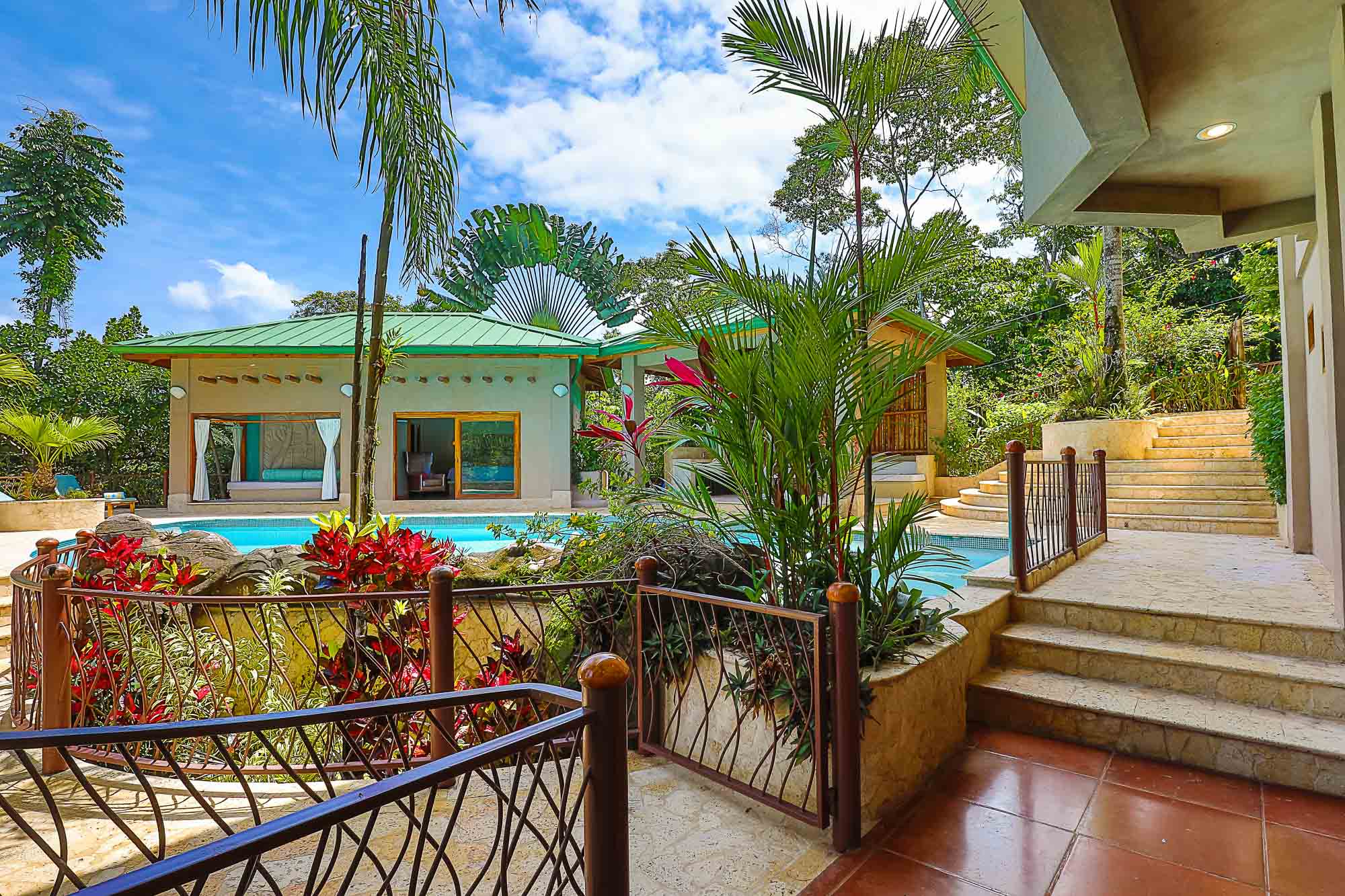 An inviting swimming pool is integrated with a tropical garden and tiered stone pathways, connecting to a bedroom pavilion that opens directly to the outdoor space under a bright sky.