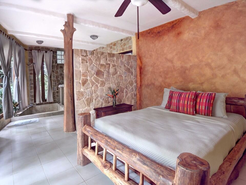 This bedroom features a rustic log bed, natural stone walls, and an integrated bath area with a tub positioned by windows overlooking tropical greenery.