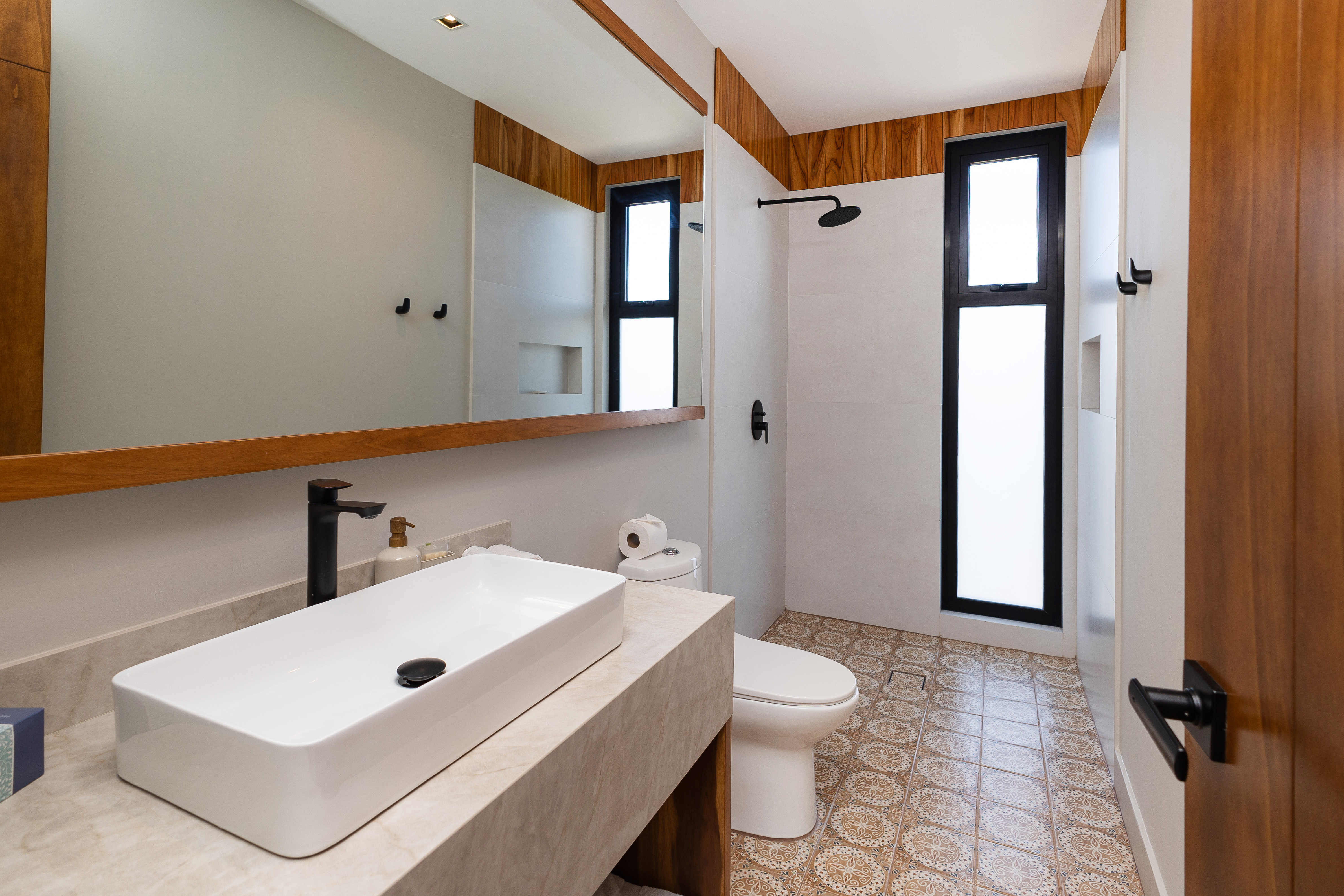 A contemporary bathroom features a rectangular basin on a light vanity, a large mirror framed in wood, a walk-in shower with matte black fixtures, and patterned floor tiles, brightened by natural light from tall vertical windows.