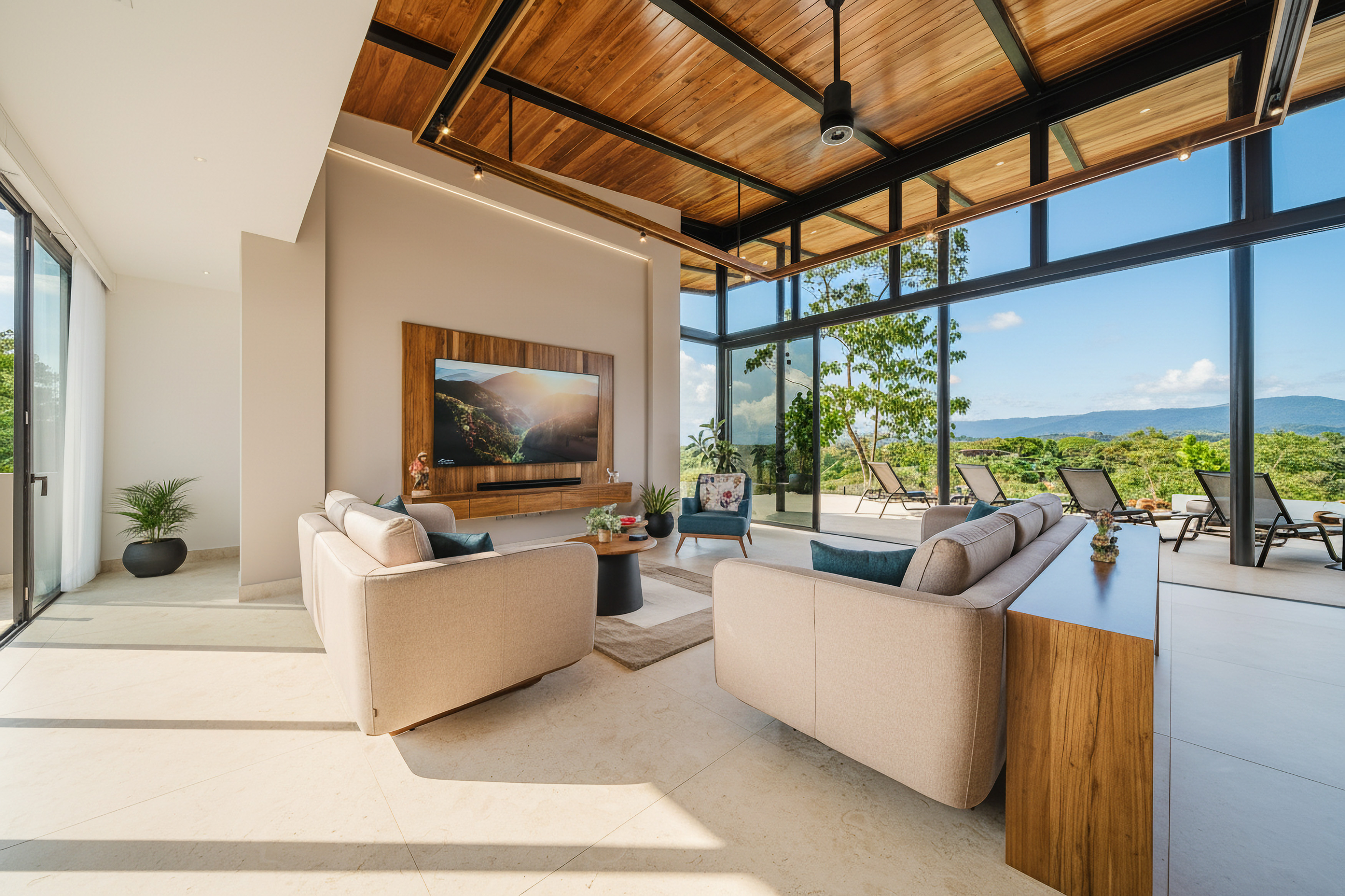 A bright living room features light-toned sofas, a wooden slatted ceiling with a fan, and floor-to-ceiling glass walls providing panoramic views of green mountains and opening to an outdoor terrace.