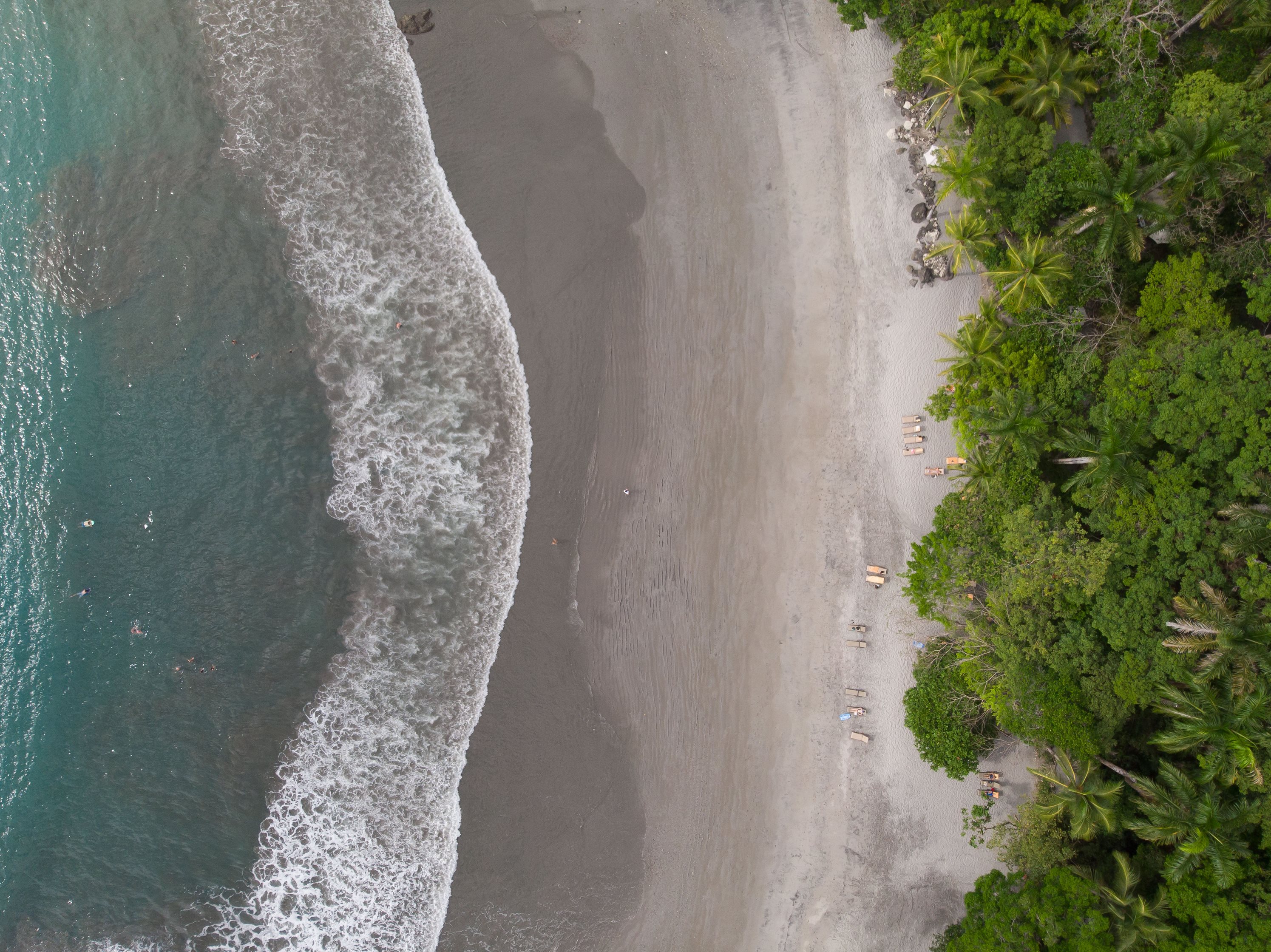 An aerial view captures the turquoise Pacific Ocean with breaking waves, a wide sandy beach, and lush tropical jungle extending to the shoreline.