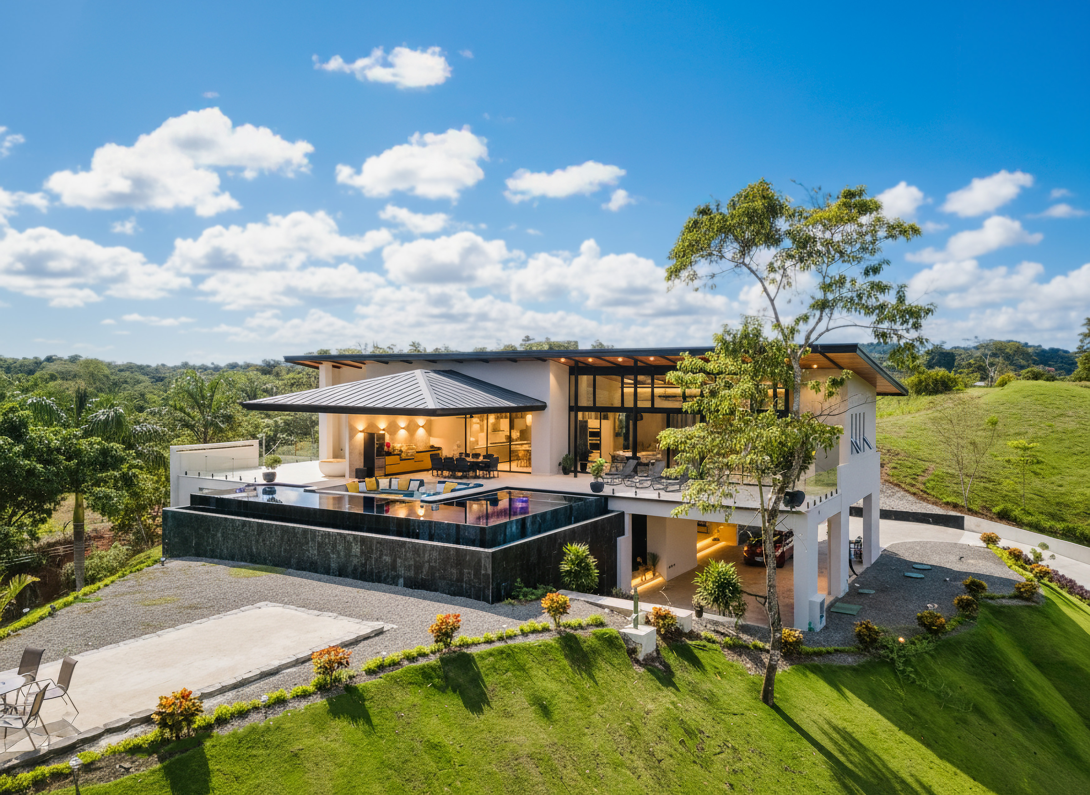 A modern multi-level villa with an infinity pool and expansive glass walls integrates with the surrounding tropical landscape under an open sky.