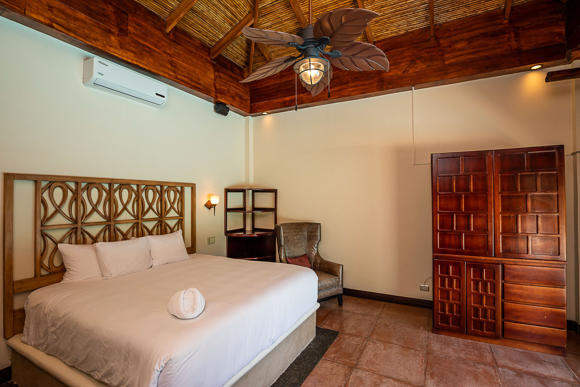 This bedroom features a high vaulted ceiling with exposed wood beams and natural woven panels, a large bed with a decorative wooden headboard, a spacious wooden wardrobe, and a tropical leaf-bladed ceiling fan, offering a comfortable and rustic ambiance.