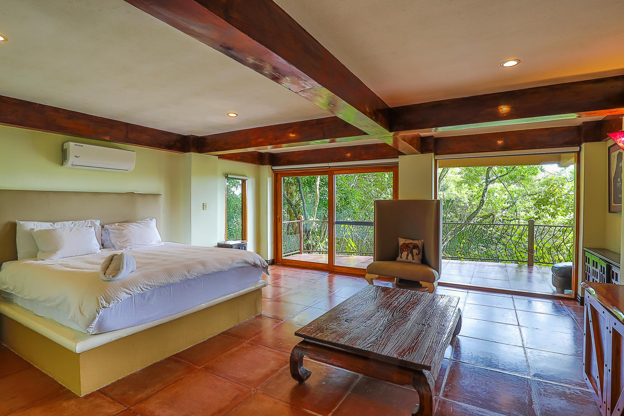 This bedroom features a large bed, exposed wooden ceiling beams, and sliding glass doors opening to a terrace that provides extensive rainforest views and natural light.
