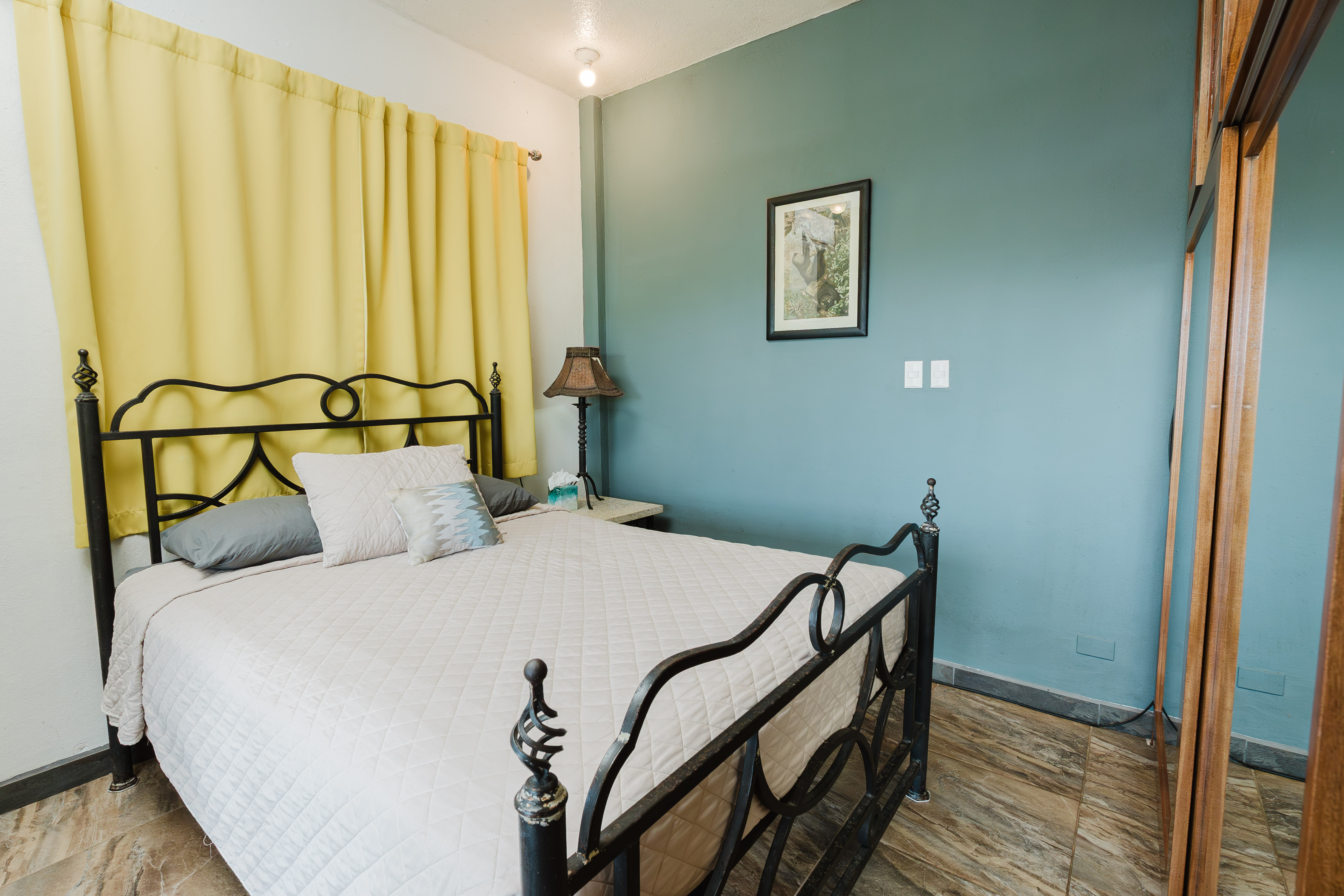 This bedroom features a black wrought iron bed frame, a distinct teal accent wall, and light-colored tiled floors, with yellow curtains suggesting natural light from a window.