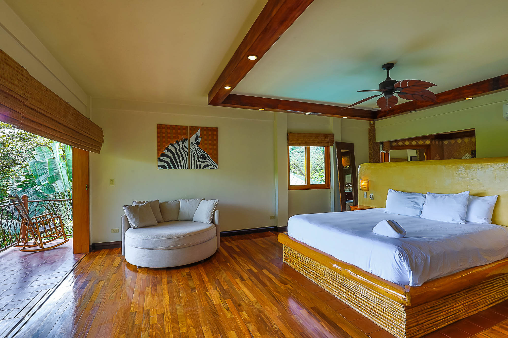A bedroom features natural wood floors, a beamed ceiling, a large bed, a round chaise lounge, and a private balcony overlooking a tropical rainforest.