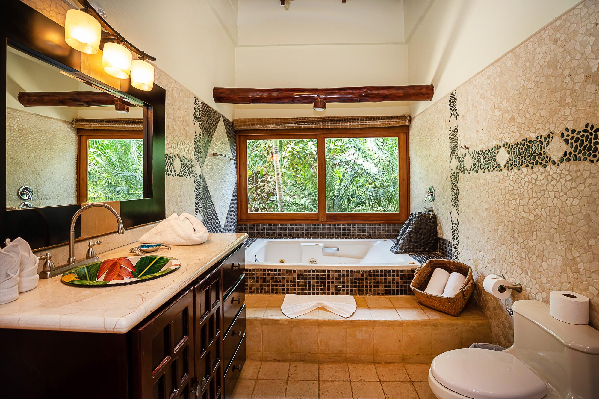 This bathroom features a deep soaking tub positioned under a wide window offering views of the tropical rainforest, a large vanity with a single sink, and walls adorned with varied stone mosaic tiles and a natural wood beam.