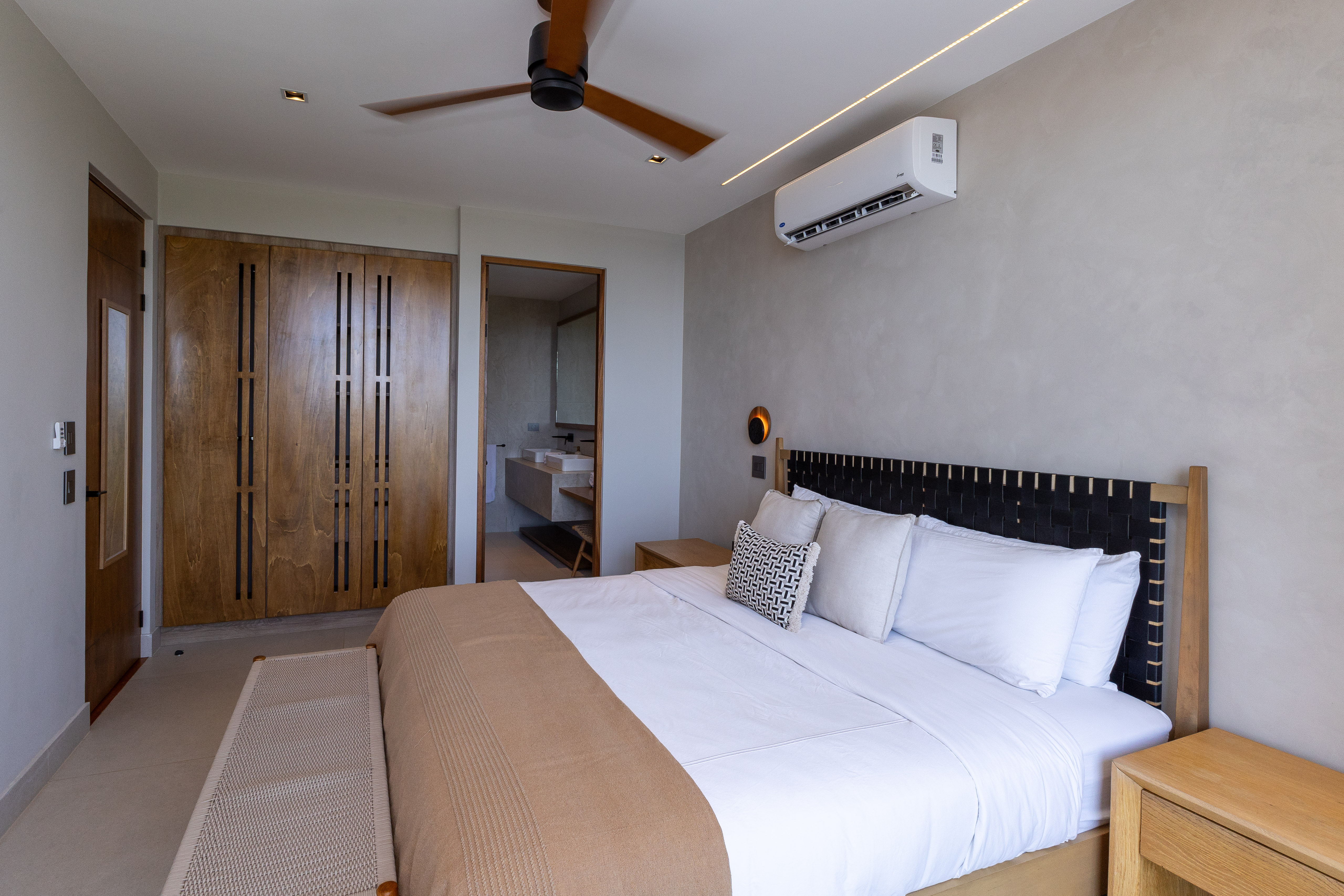 This contemporary bedroom features a large bed with a woven headboard, built-in wooden closets, a wall-mounted air conditioning unit, a ceiling fan, and an en-suite bathroom visible through an open doorway.