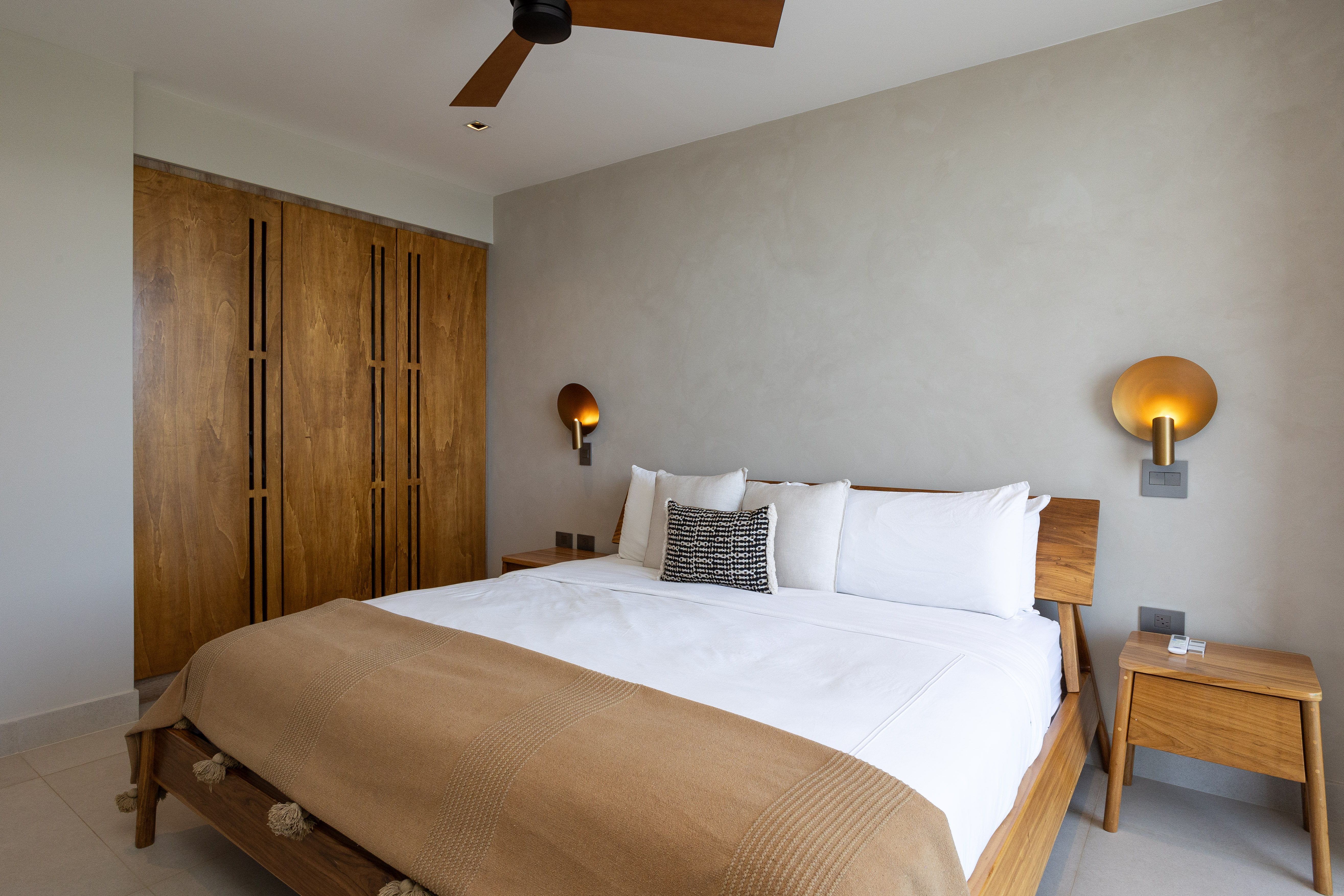 A contemporary bedroom features a wooden bed frame, matching nightstands, and built-in wardrobes, complemented by light gray walls and a wooden ceiling fan overhead.