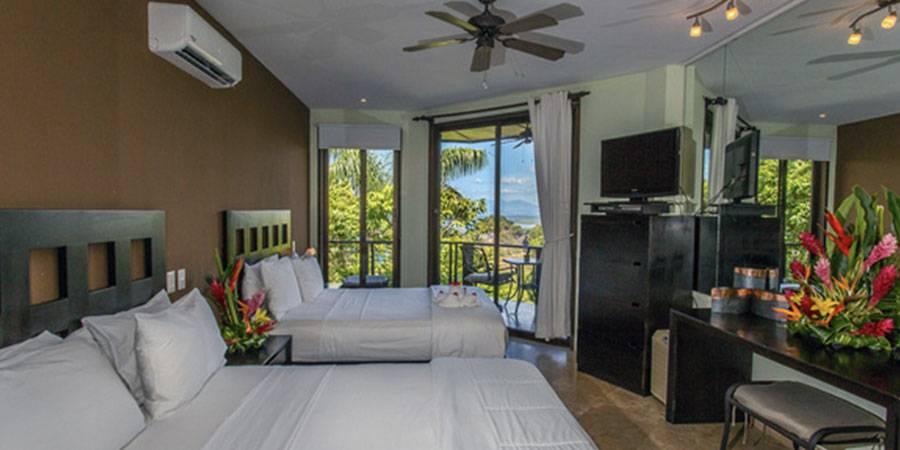 This bright bedroom features two queen beds, a ceiling fan, and large glass doors leading to a private balcony with tropical jungle and ocean views.