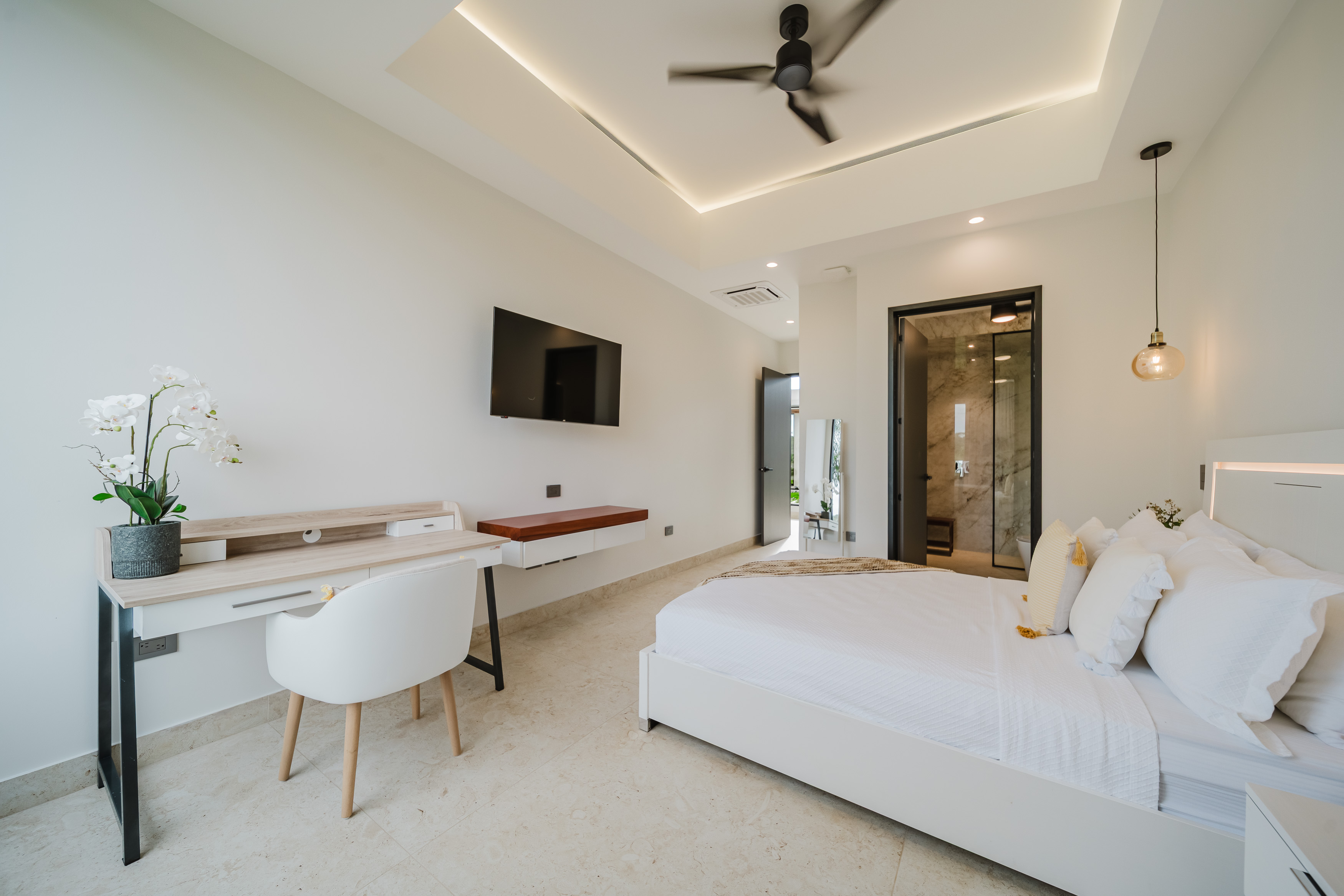 A bright bedroom features a modern bed with integrated headboard lighting, a desk area, a wall-mounted television, and direct access to an outdoor area and a private bathroom with a visible marble shower.