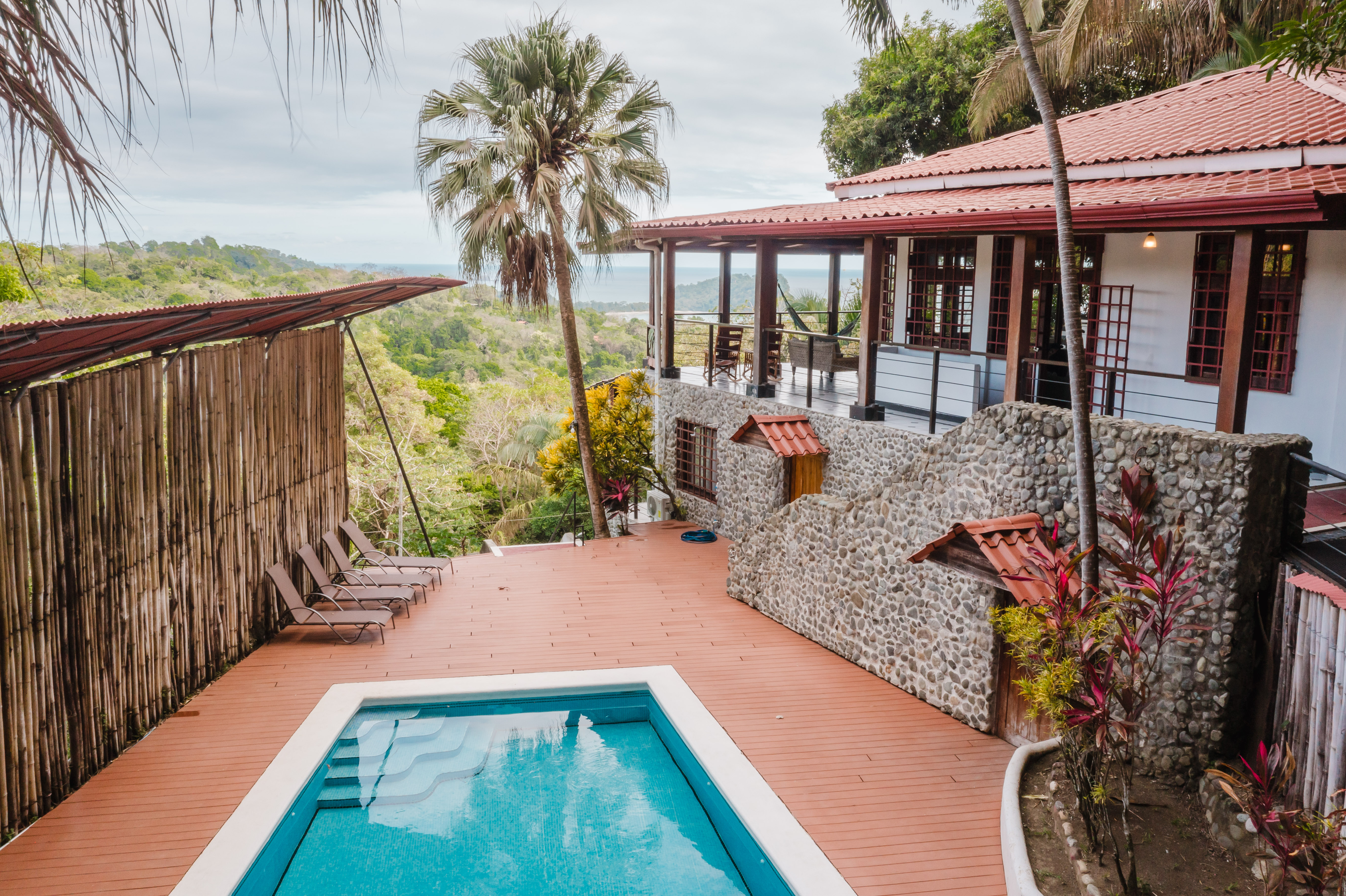 A private tiled pool with steps is surrounded by a spacious wooden deck, leading to an elevated covered terrace offering tropical jungle and ocean views.