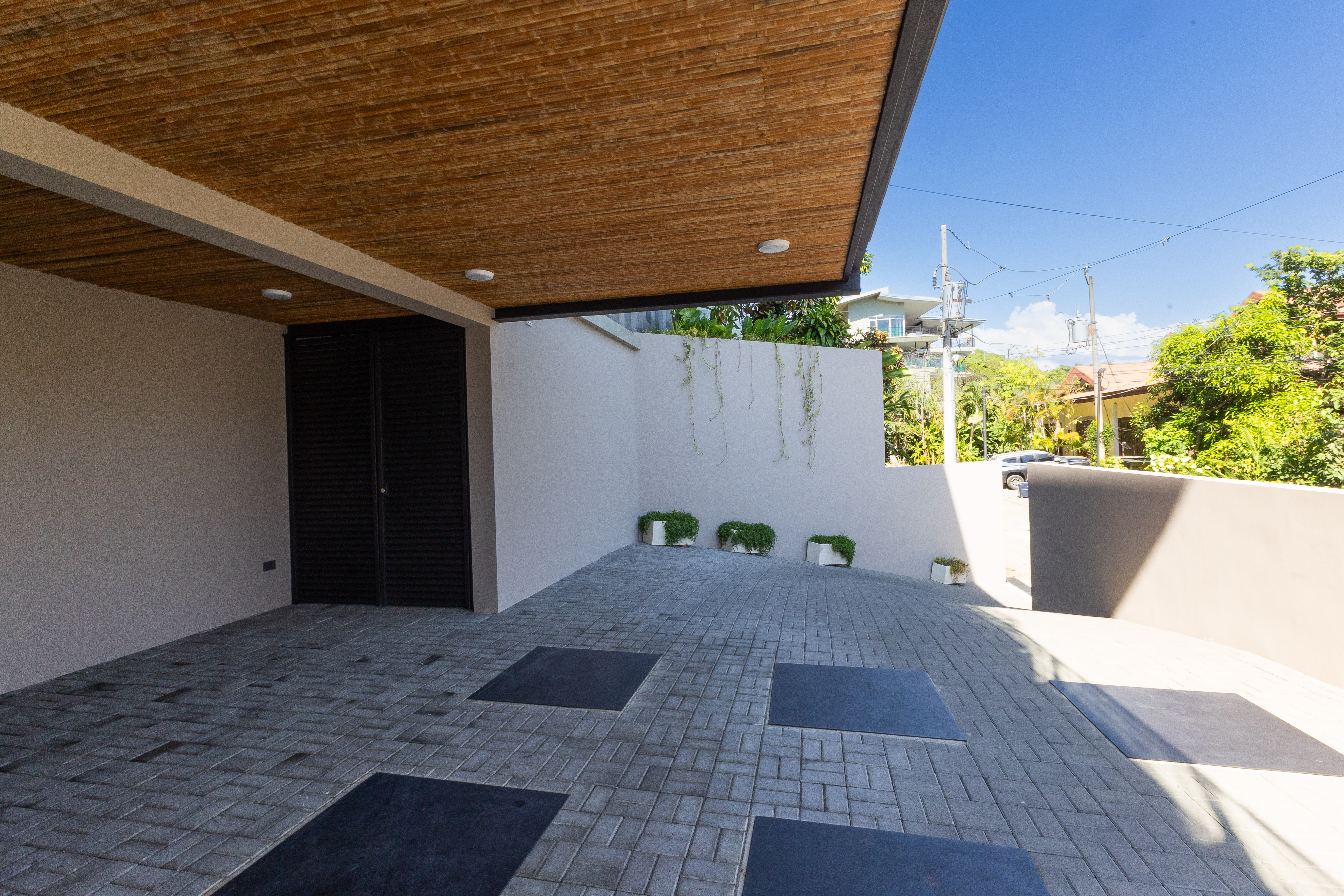 A modern covered carport features a bamboo-paneled ceiling and paved drive, with bright natural light revealing tropical foliage beyond.