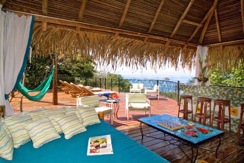 The open-air Sky Deck, covered by a thatched roof, features a daybed, lounge chairs, and a bar, all bathed in natural light and overlooking the ocean and dense jungle canopy.