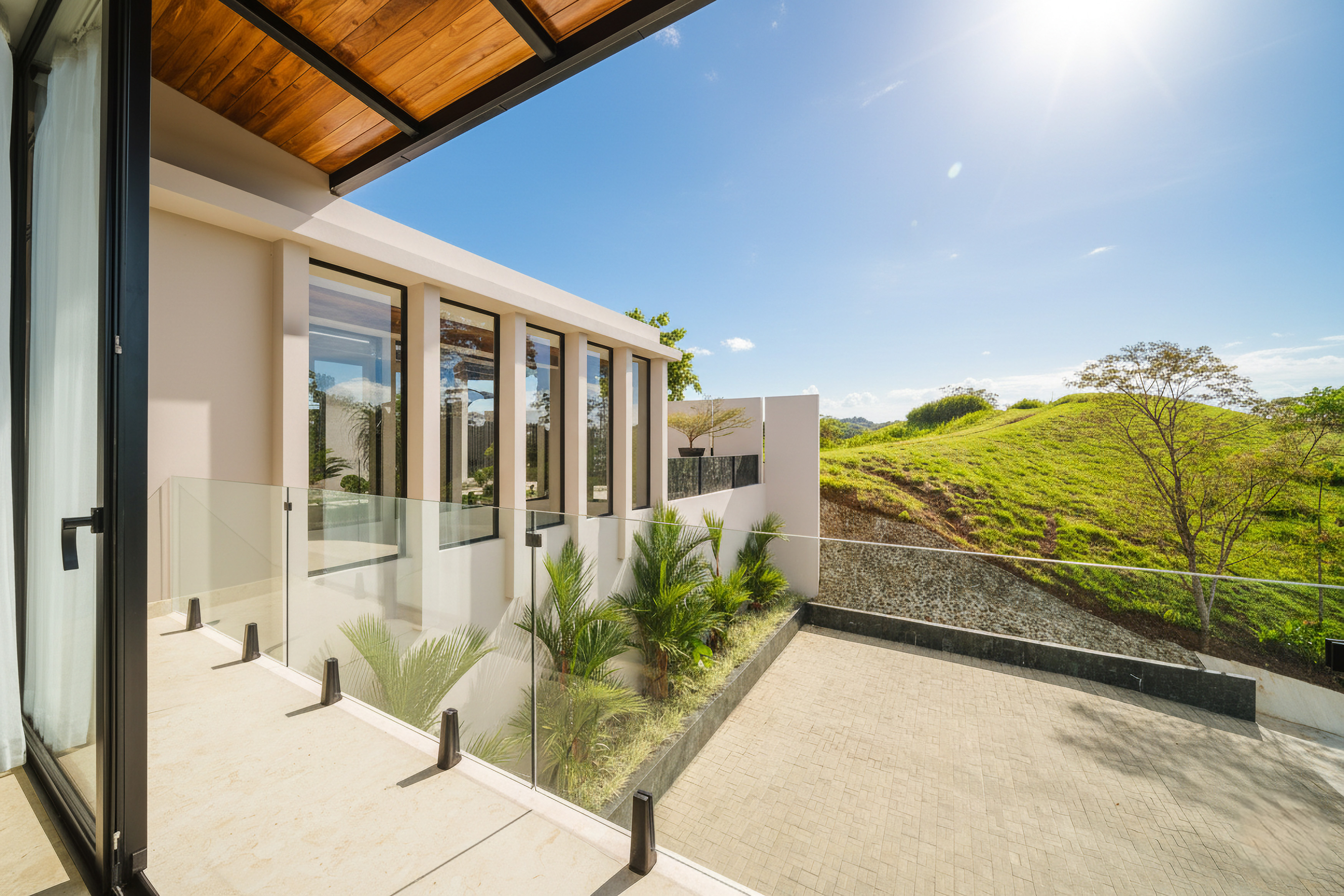 A covered outdoor terrace features a wooden ceiling and glass railing, offering views of a modern building facade, a paved patio, and sunlit green hills under a clear blue sky.