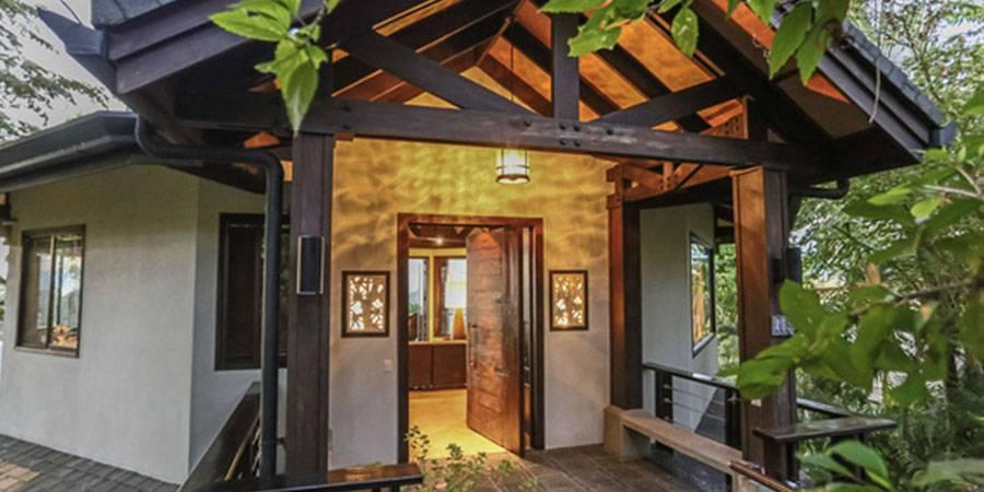 The villa's covered entrance showcases dark wood beams, a natural wood ceiling, and a large open wooden door revealing a warm interior, all set amidst tropical greenery.