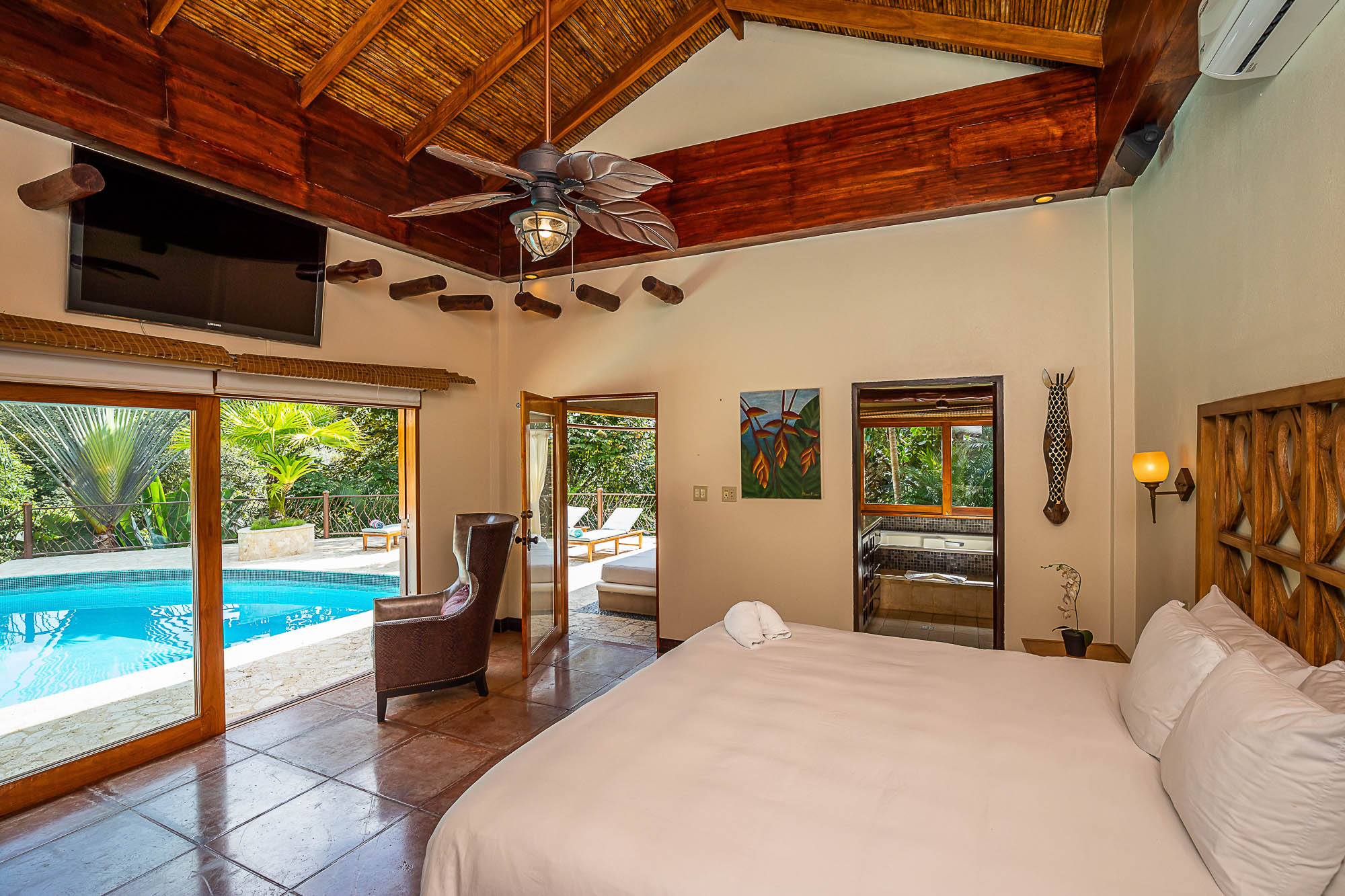 A spacious bedroom features a vaulted ceiling with dark wood beams and bamboo, terracotta tile floors, direct access to a swimming pool, and an adjacent terrace, offering views of tropical foliage and ample natural light.
