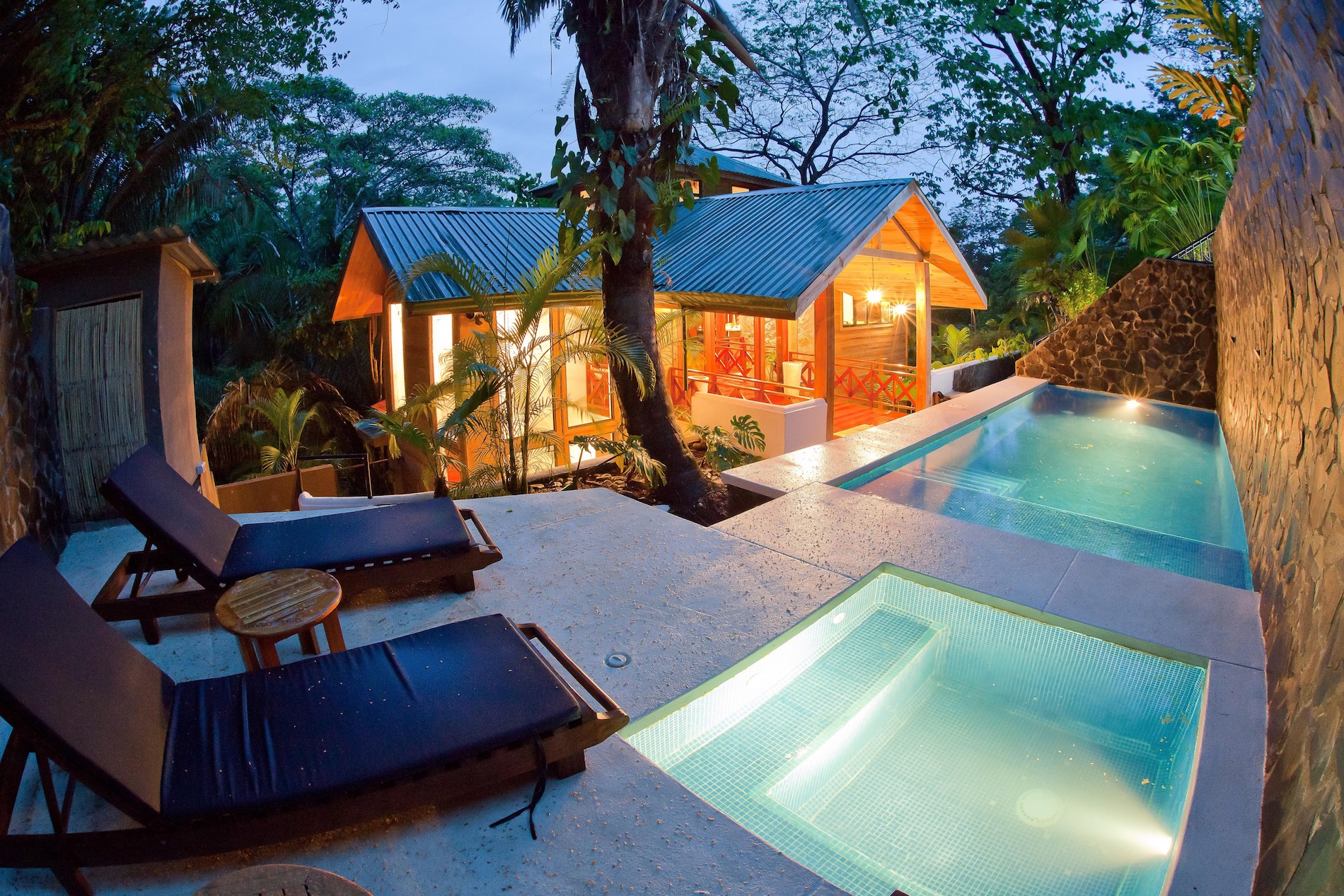 An illuminated private plunge pool and separate jacuzzi sit on a stone terrace overlooking a warm-lit wooden villa and lush jungle foliage at twilight.