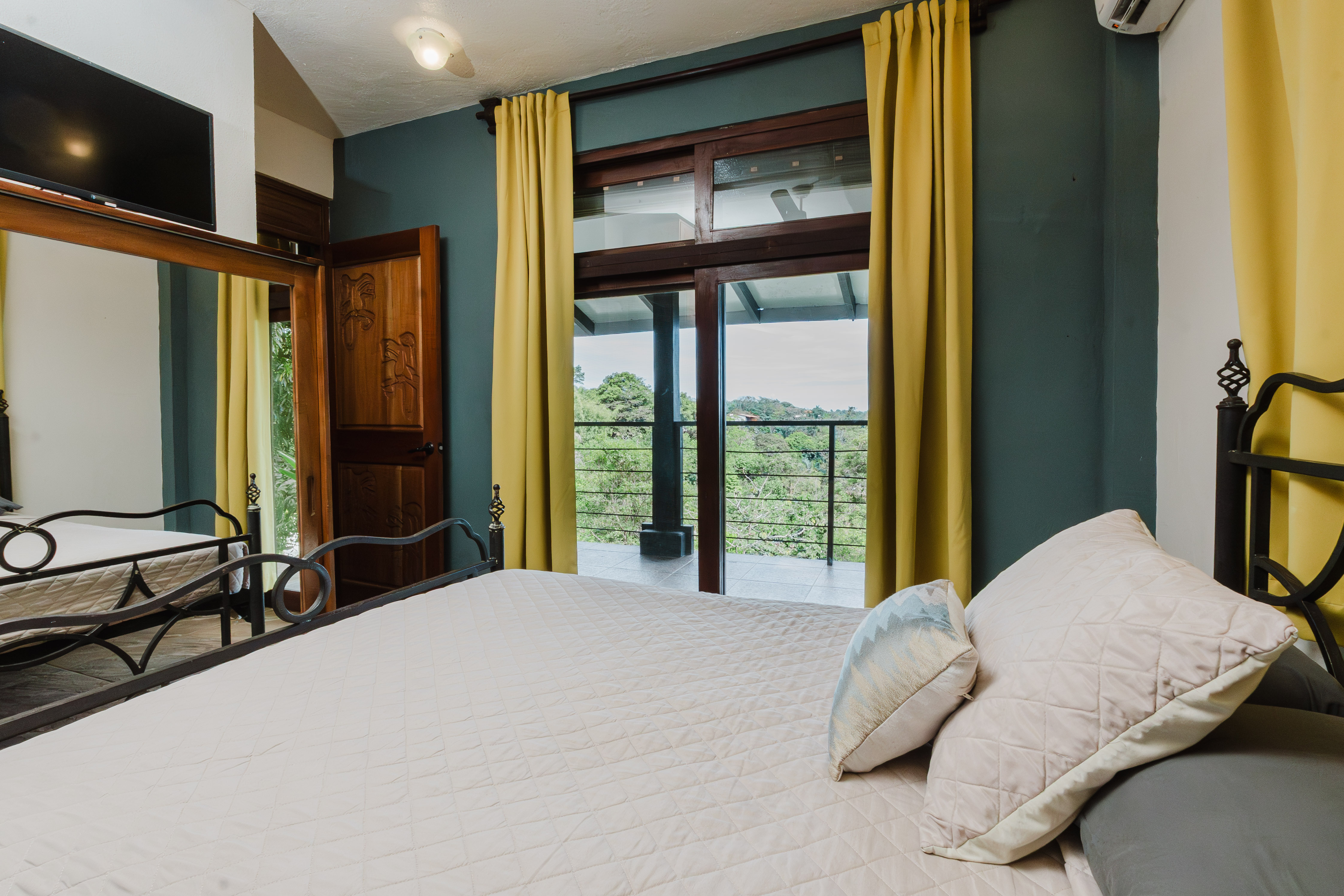 This air-conditioned bedroom features a bed, a carved wooden door, and large glass doors that open to a private balcony overlooking a tropical landscape, providing ample natural light.