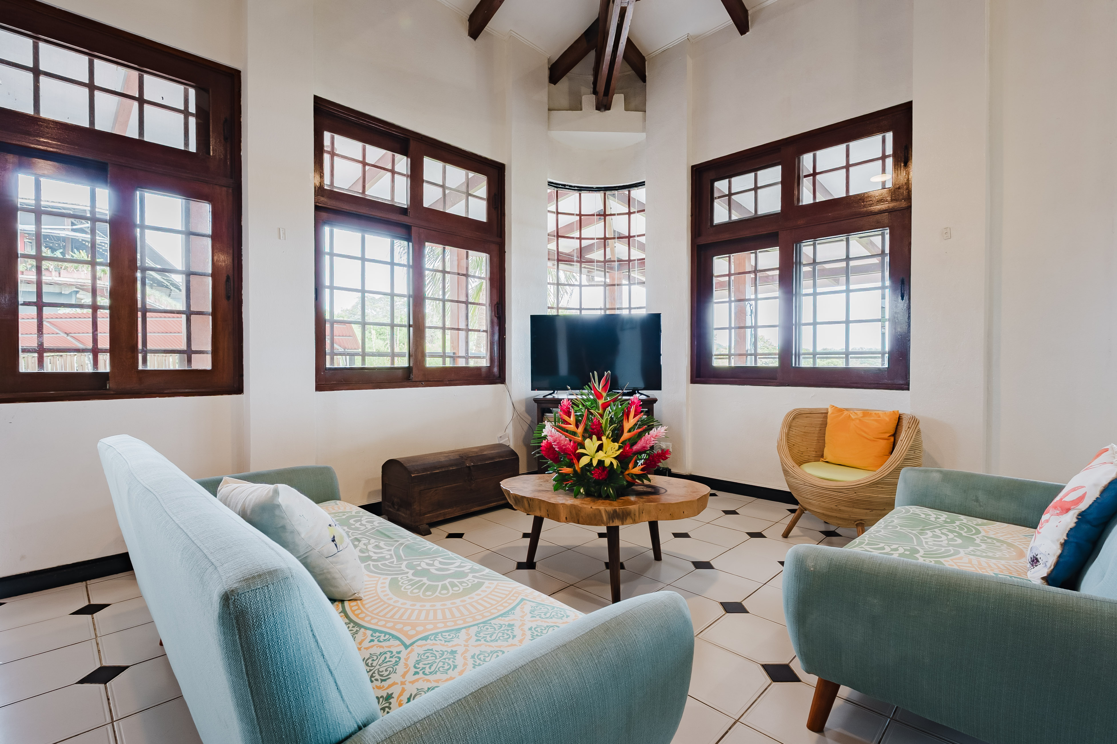A bright living room features a high vaulted ceiling with exposed wooden beams, multiple large grid-paned windows offering natural light and views of tropical greenery, and seating on light blue upholstered furniture.
