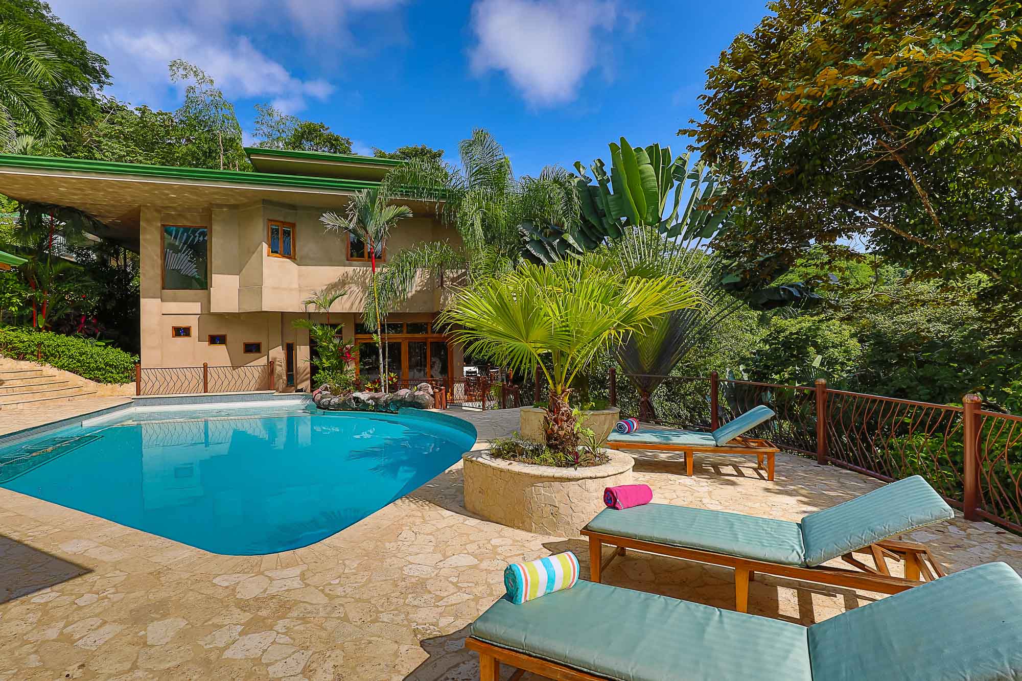 A large outdoor swimming pool is set within a stone patio featuring sun loungers, with a multi-story villa and dense tropical foliage creating a secluded environment under open skies.
