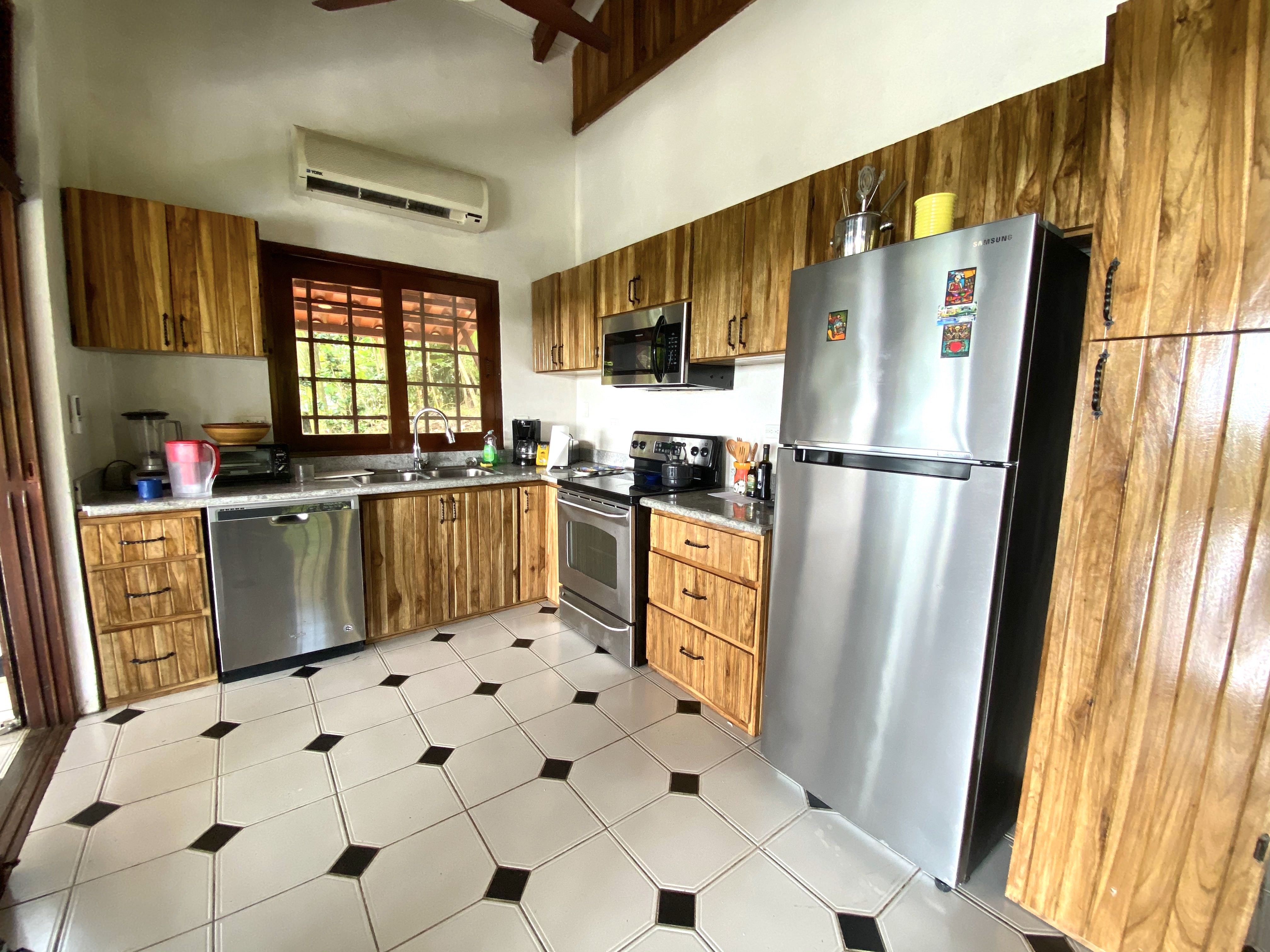 This kitchen features rich natural wood cabinetry, a patterned tiled floor, stainless steel appliances, and a window above the sink offering views of tropical foliage and natural light, complemented by an overhead air conditioning unit.