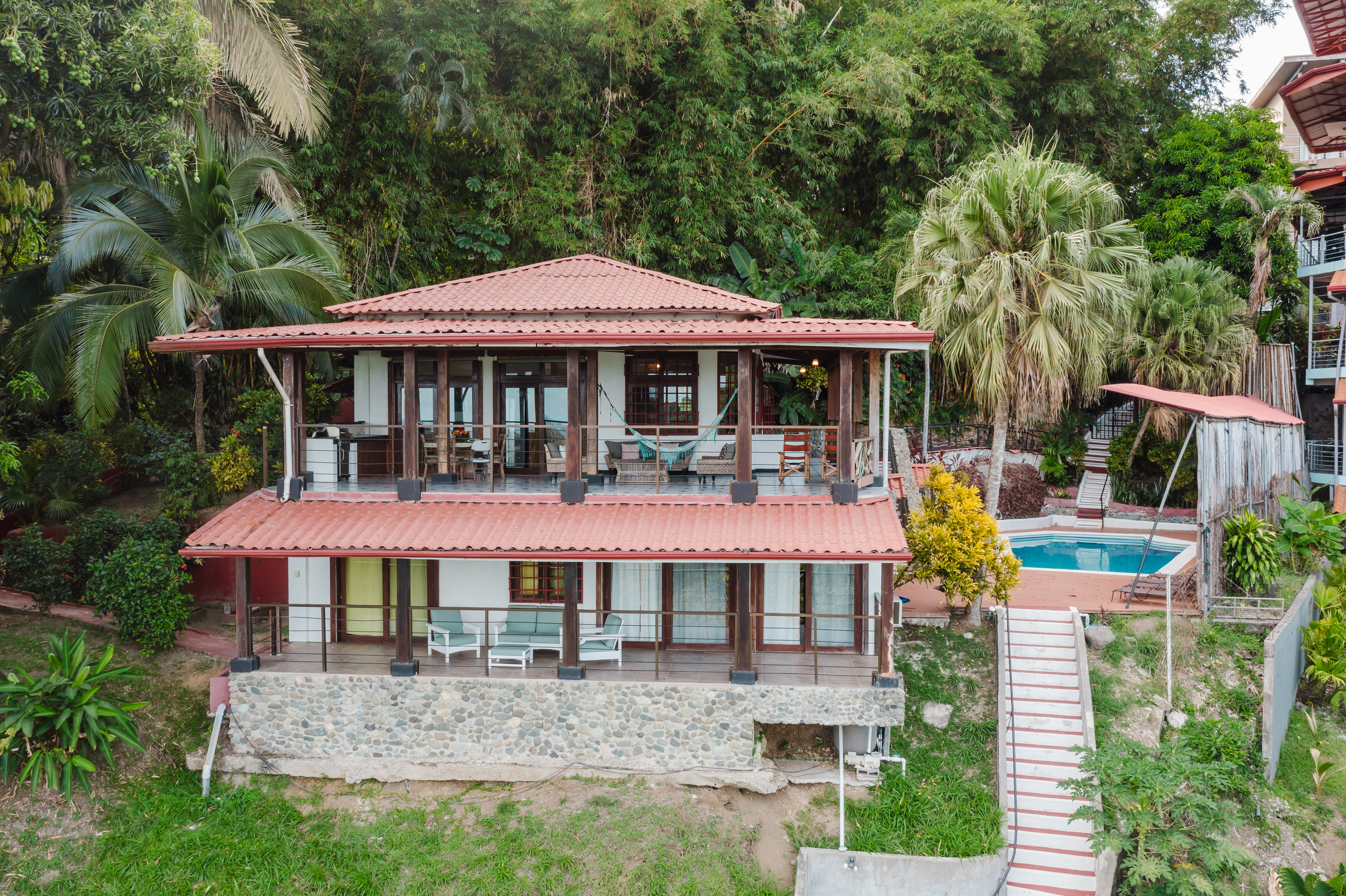 A two-story villa with red tile roofs features open-air terraces on both levels and a private pool, surrounded by dense tropical greenery.