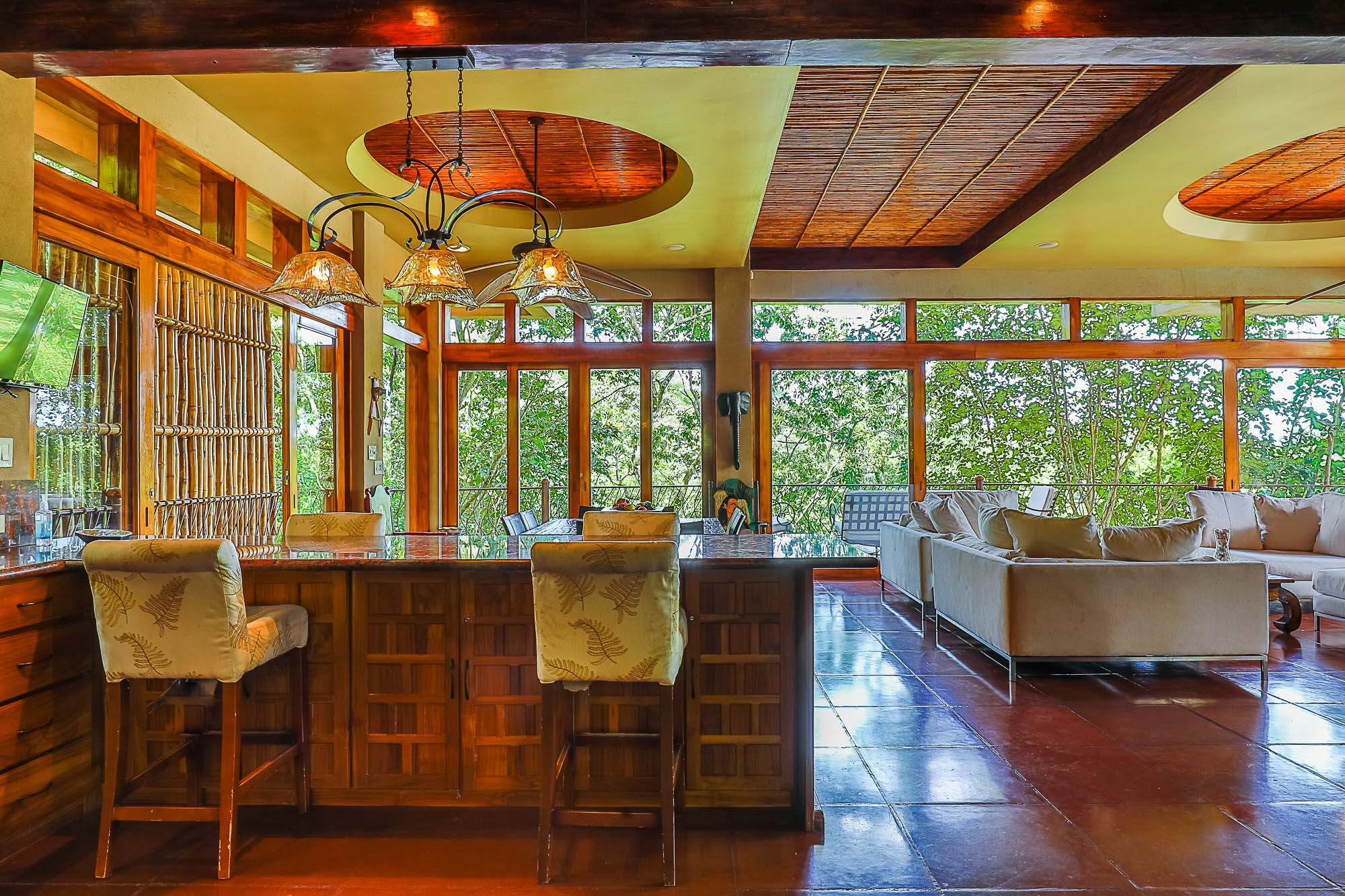 The open-plan living space features a wooden bar with stools and a lounging area with sofas, offering natural light and expansive views of the surrounding rainforest through large windows.