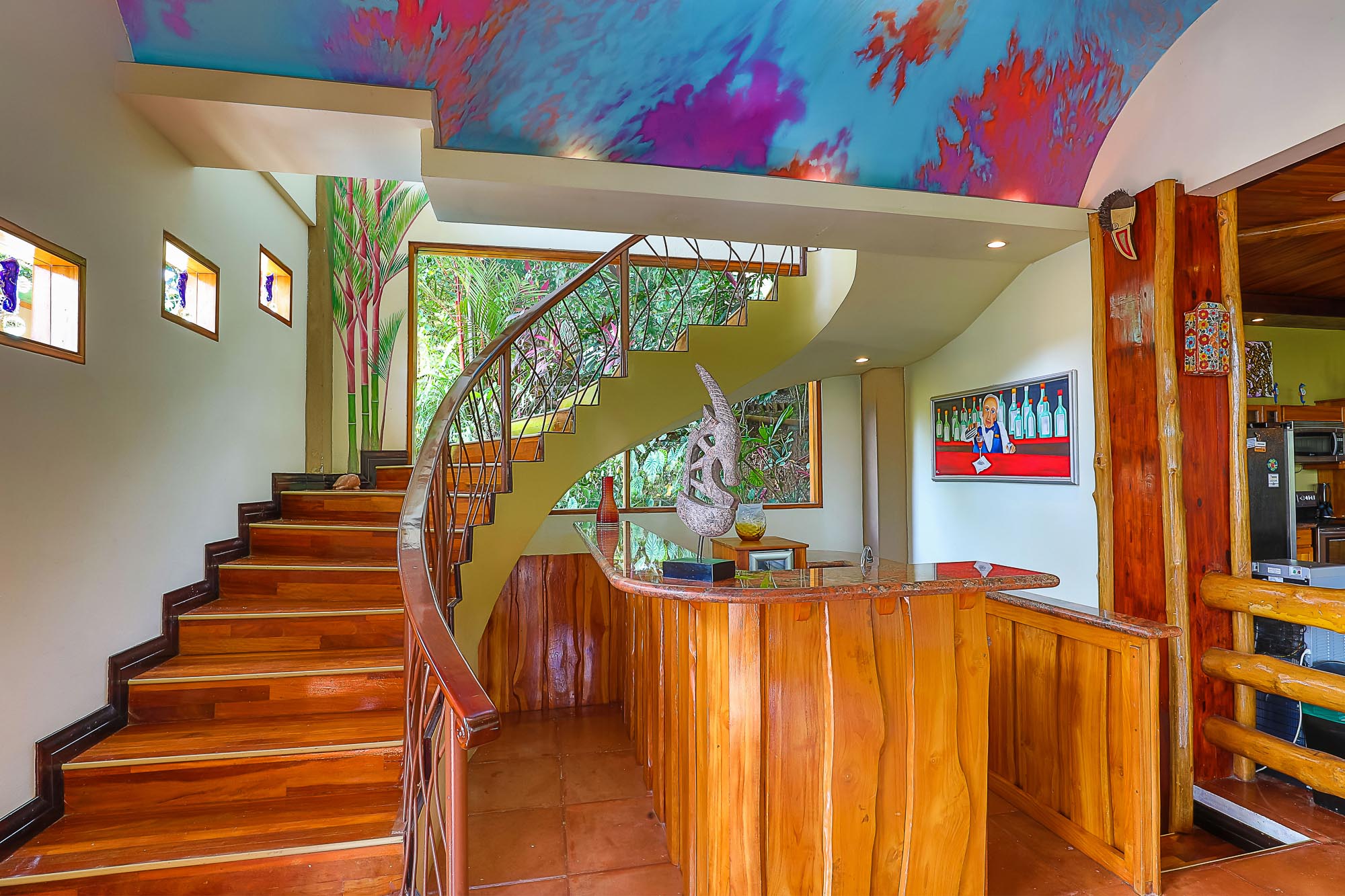 This vibrant entry space features a curving wooden staircase with an ornate metal railing, a natural wood bar, and large windows opening to lush tropical views, all beneath a colorful abstract ceiling mural.