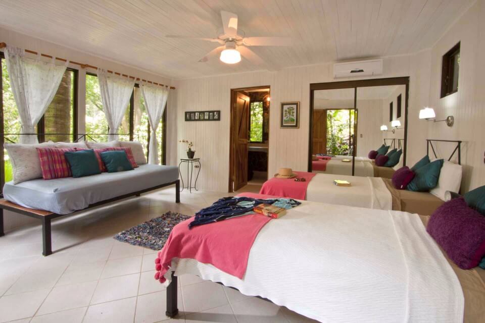 This airy bedroom features white paneled walls, two single beds, a daybed, and receives ample natural light and lush jungle views through large windows.