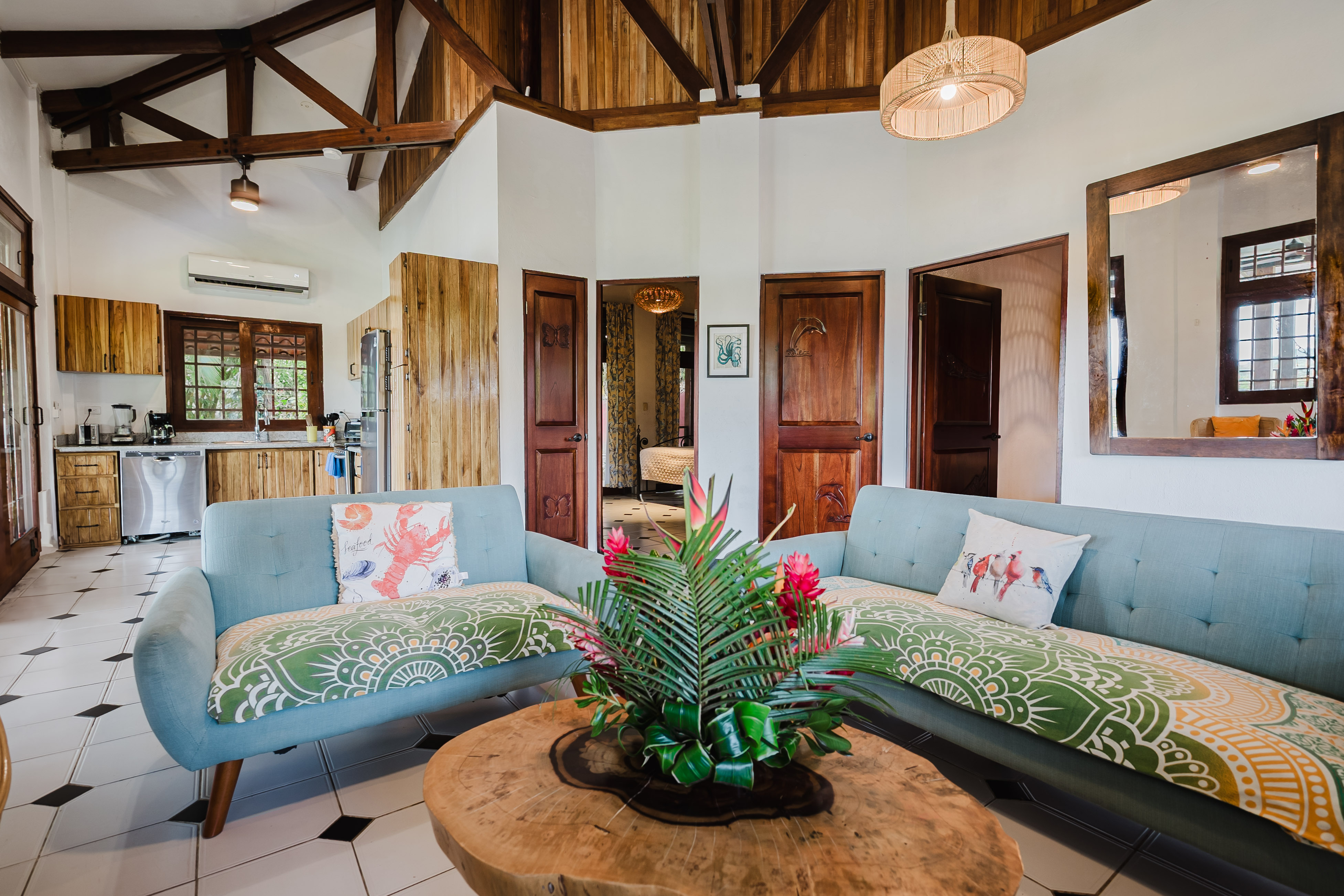 This bright, open-plan living area and kitchen features a high ceiling with exposed wood beams and paneling, light tiled floors, two sofas, and natural wood cabinetry, with views of greenery through a kitchen window.