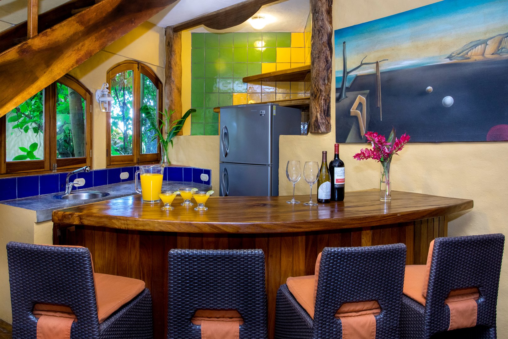 A rustic wooden wet bar with seating is naturally lit by arched windows offering views into the lush tropical jungle.