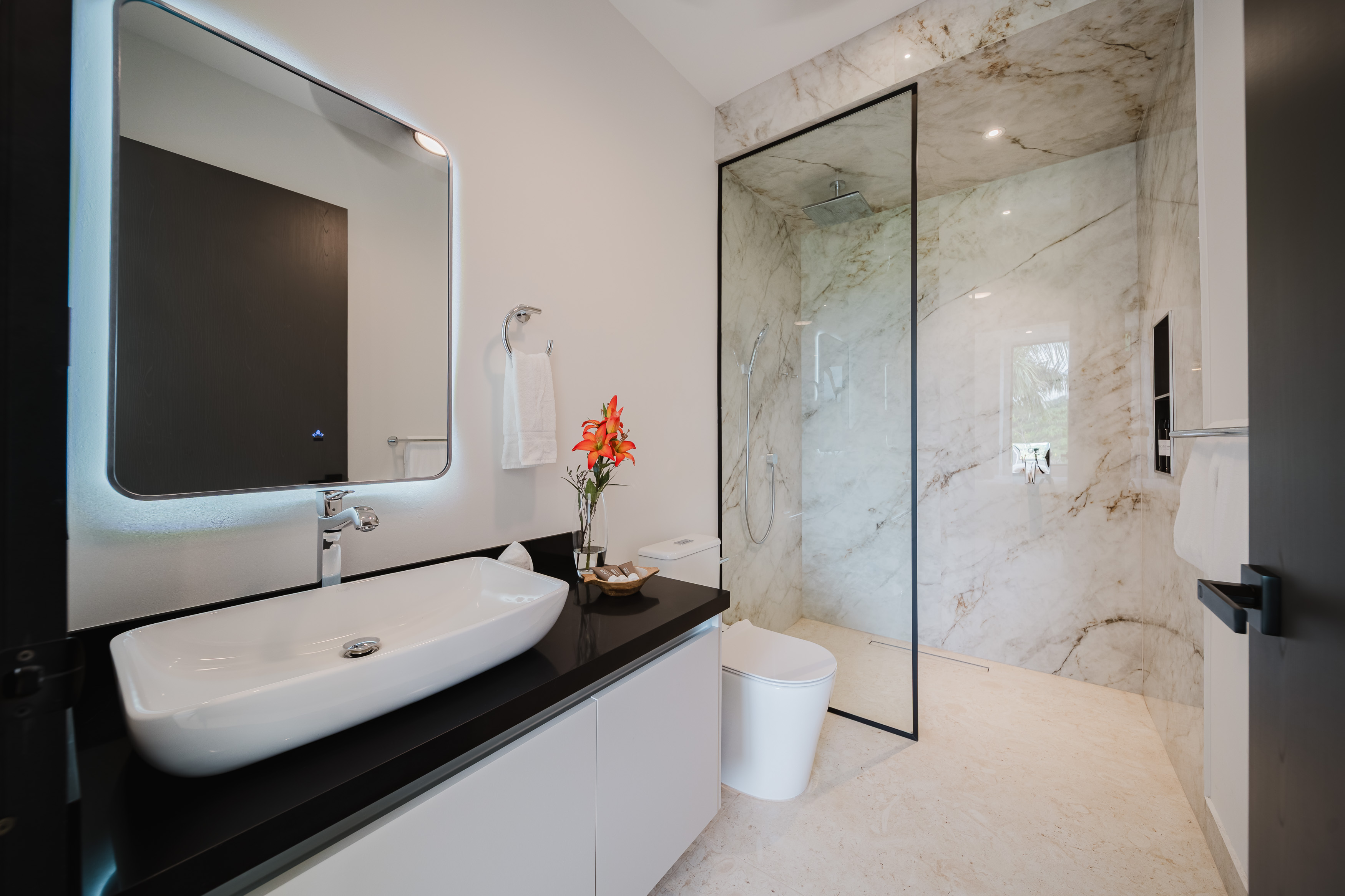 A contemporary bathroom features a floating vanity with a vessel sink, a backlit mirror, and a walk-in shower with marble-look walls and natural light from an outdoor view.