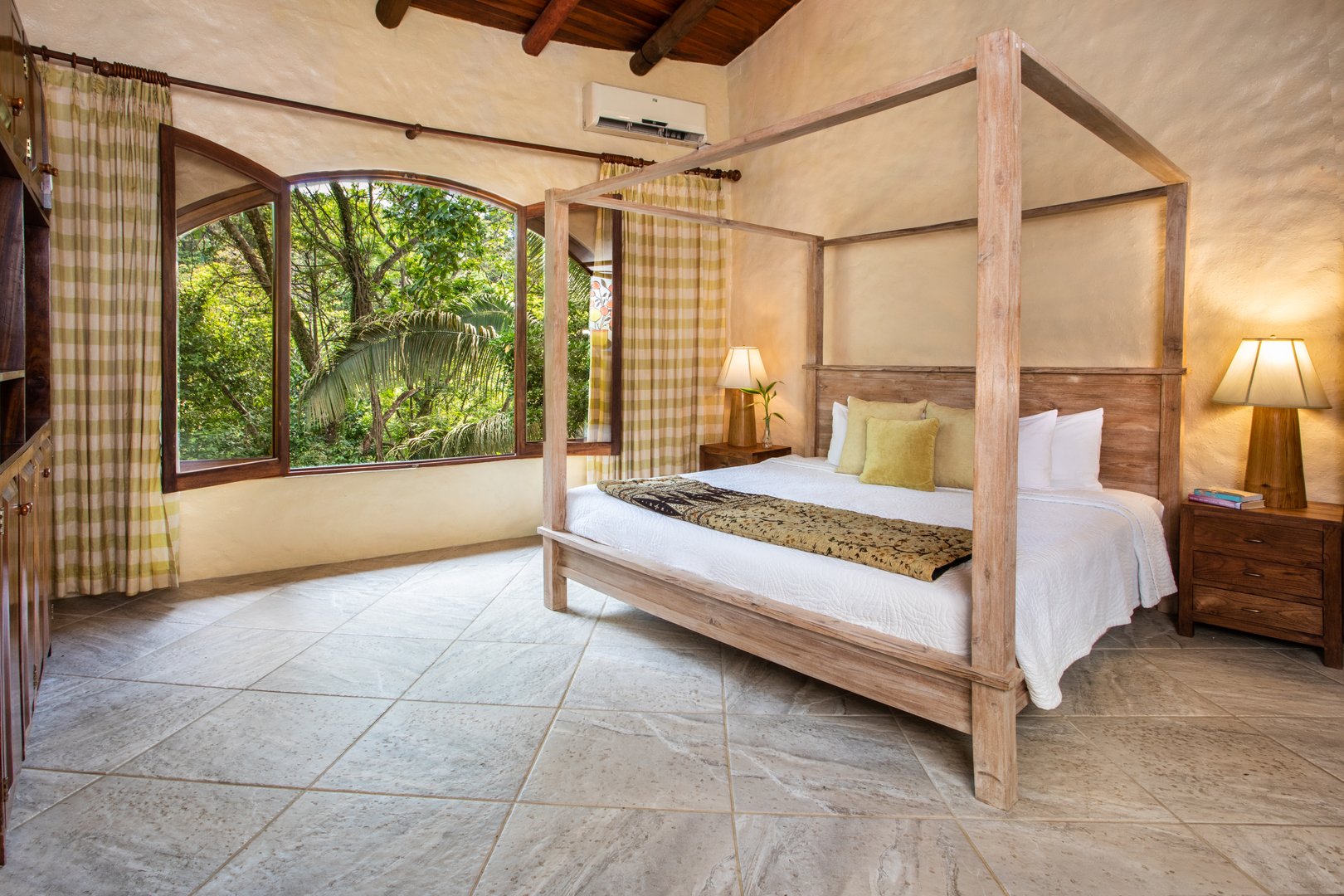 A spacious bedroom with a wooden four-poster bed and exposed ceiling beams features large arched windows that open to a lush jungle view, filling the room with natural light.
